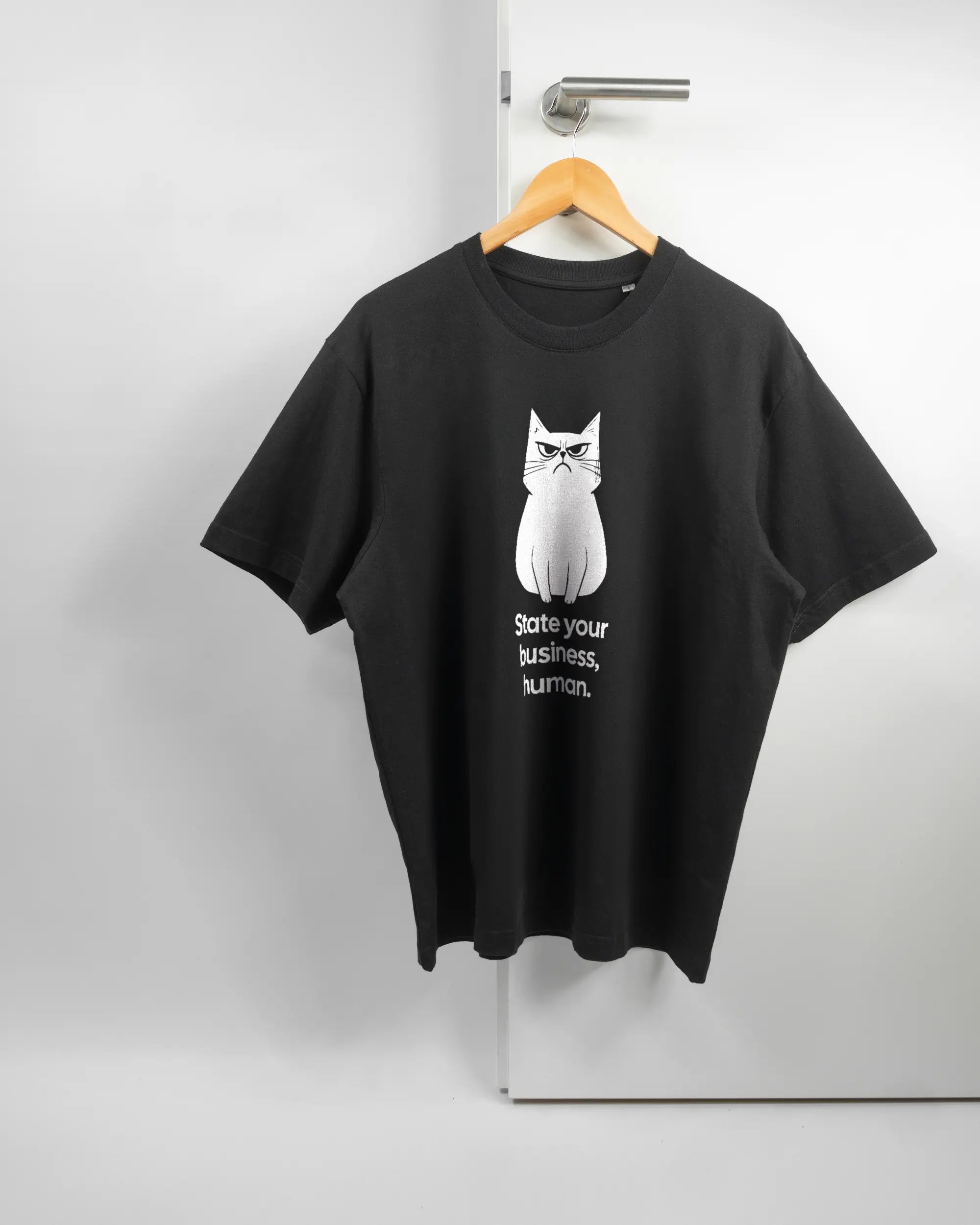 Vorderseite des Basic Unisex T-Shirt Oversized von Maondo mit dem Design Grumpy Purr in der Farbe Black und in der Größe S auf einem Bügel an der Türklinke aufgehängt