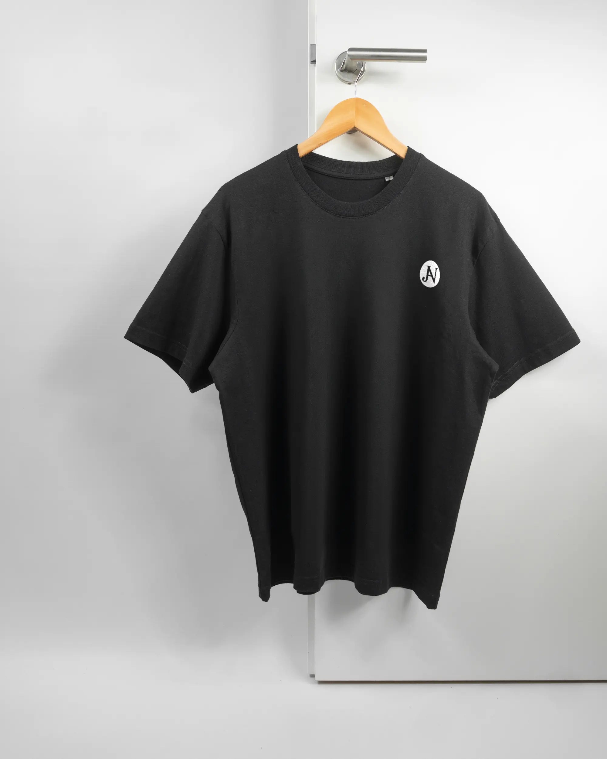 Vorderseite des Basic Unisex T-Shirt Oversized von Maondo mit dem Design JB1 in der Farbe Black und in der Größe S auf einem Bügel an der Türklinke aufgehängt