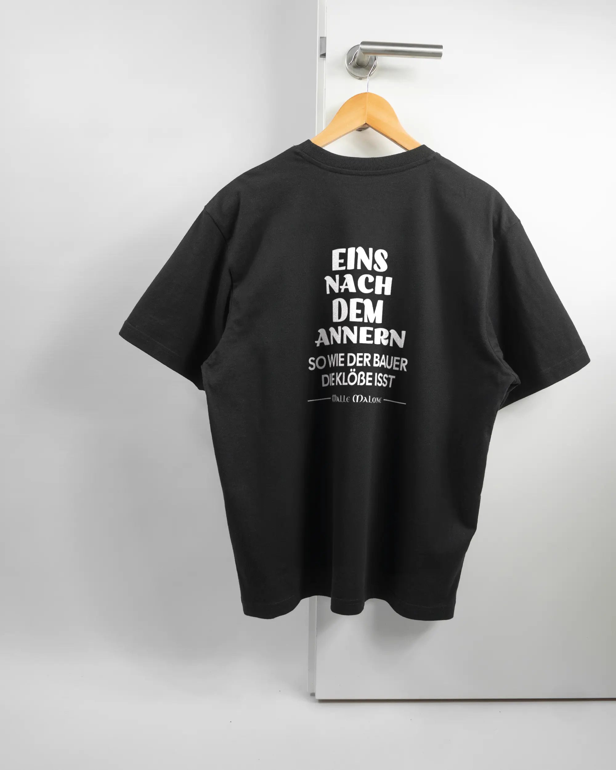 Rückseite des Basic Unisex T-Shirt Oversized von Maondo mit dem Design Eins nach dem Annern in der Farbe Black und in der Größe S auf einem Bügel an der Türklinke aufgehängt