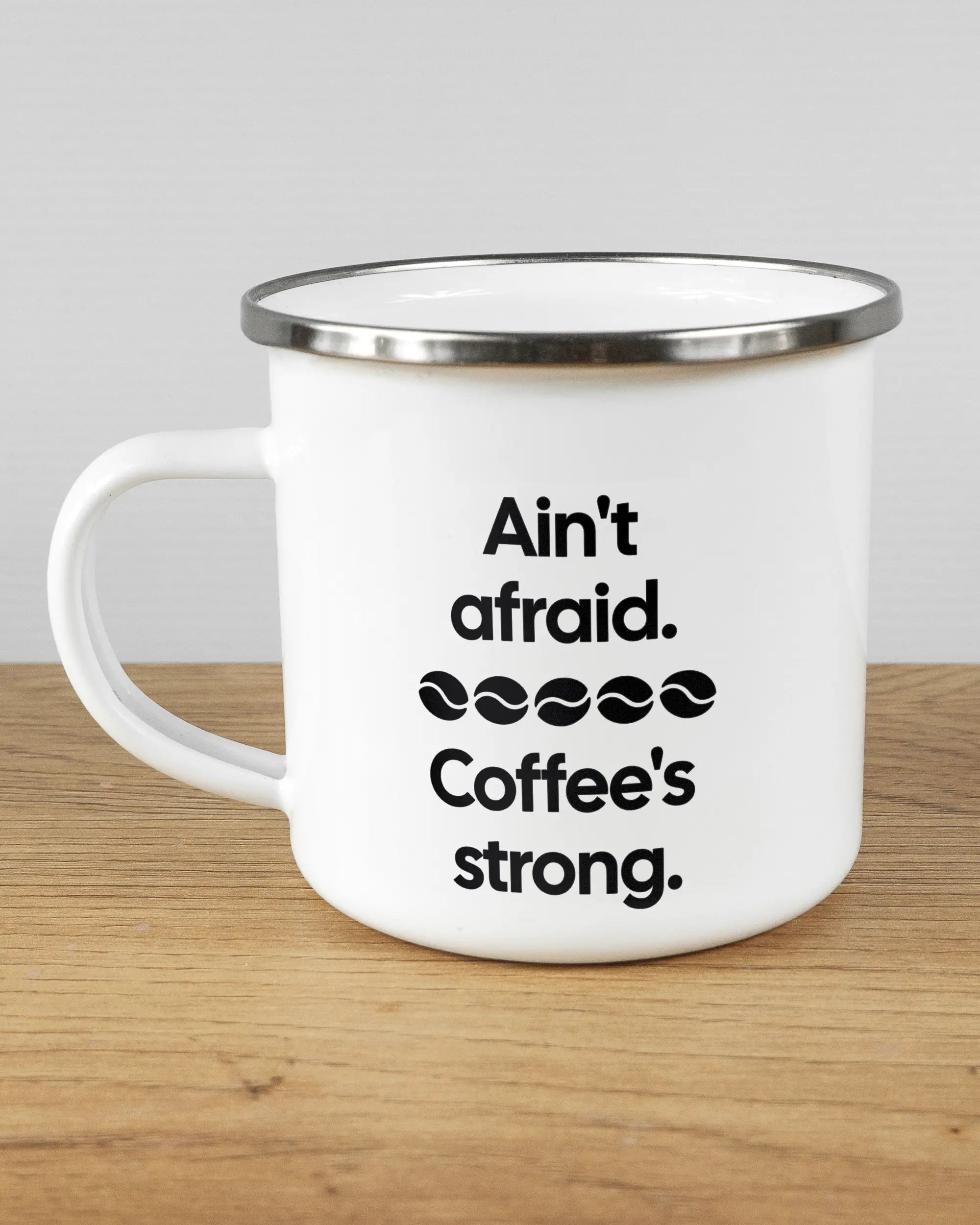 Titelbild, das die linke Seite der beidseitig bedruckten Emailletasse von Maondo mit dem Design Coffee is strong in der Farbe White-Silver auf einem Holztisch zeigt