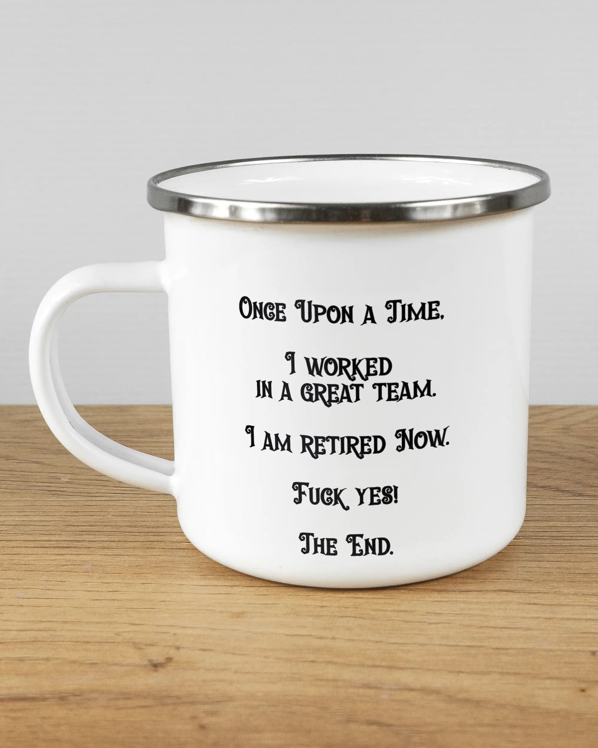 Titelbild, das die linke Seite der beidseitig bedruckten Emailletasse von Maondo mit dem Design Finally Retired in der Farbe White-Silver auf einem Holztisch zeigt