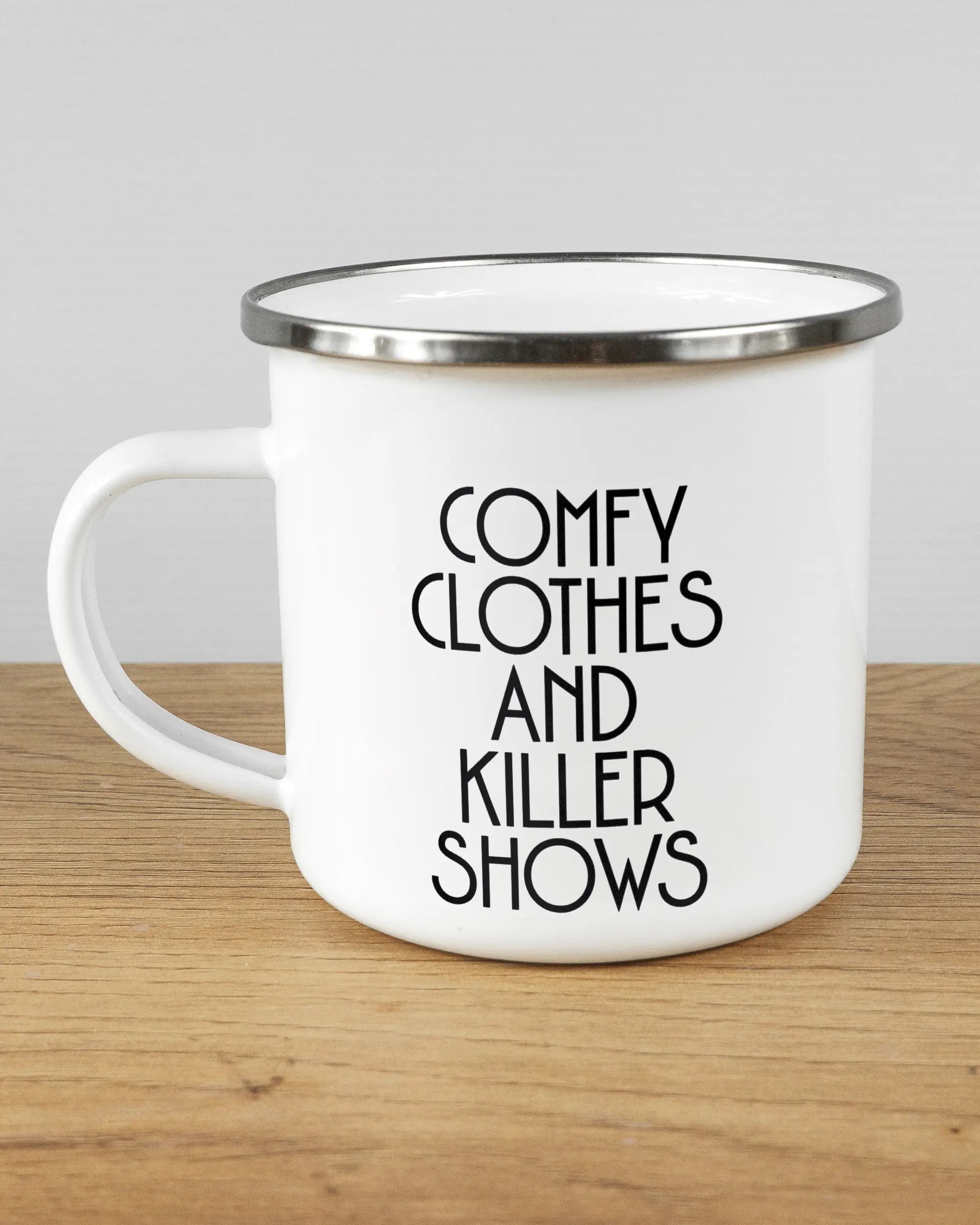 Titelbild, das die linke Seite der beidseitig bedruckten Emailletasse von Maondo mit dem Design Killer Shows in der Farbe White-Silver auf einem Holztisch zeigt