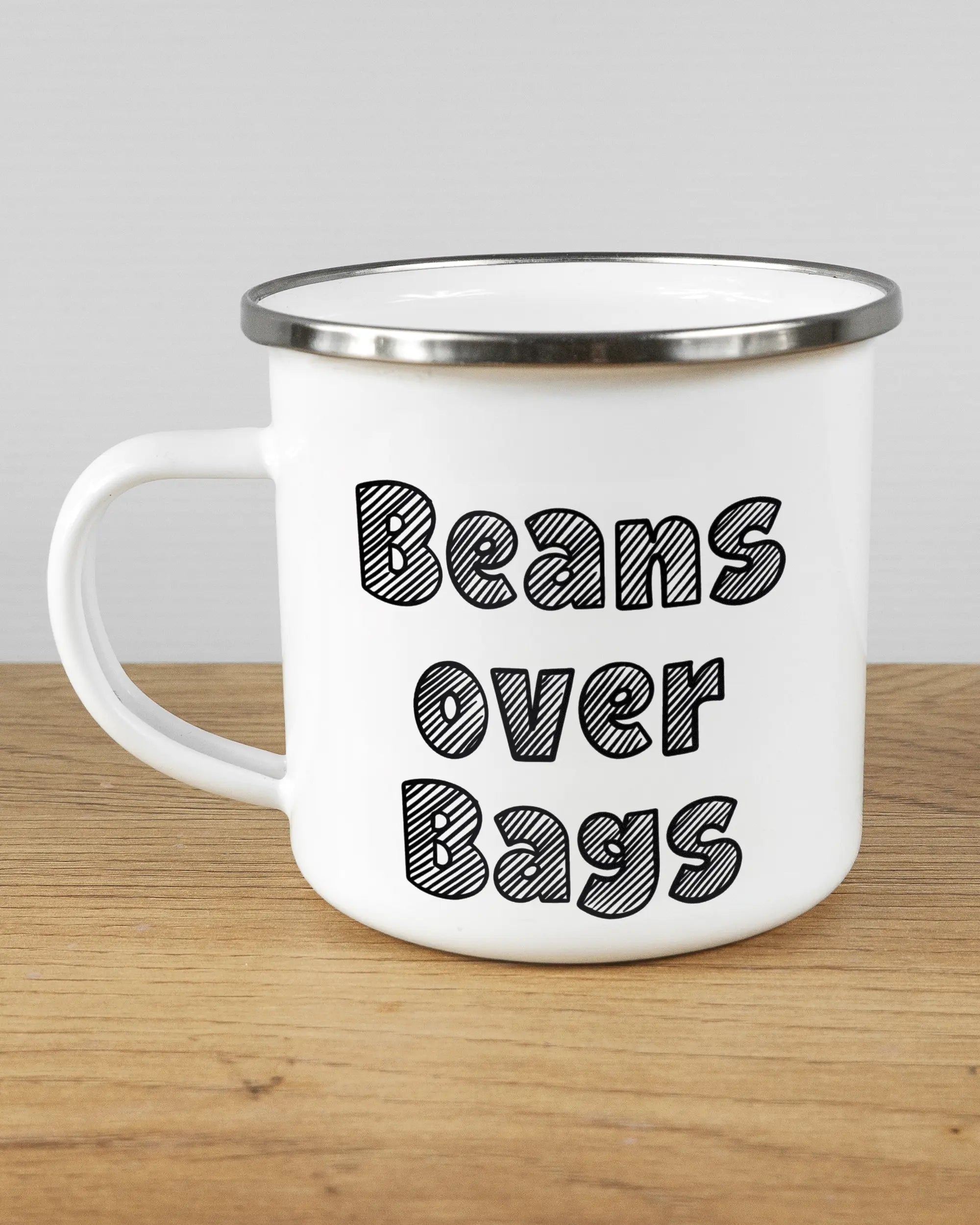 Titelbild, das die linke Seite der beidseitig bedruckten Emailletasse von Maondo mit dem Design Beans over Bags in der Farbe White-Silver auf einem Holztisch zeigt