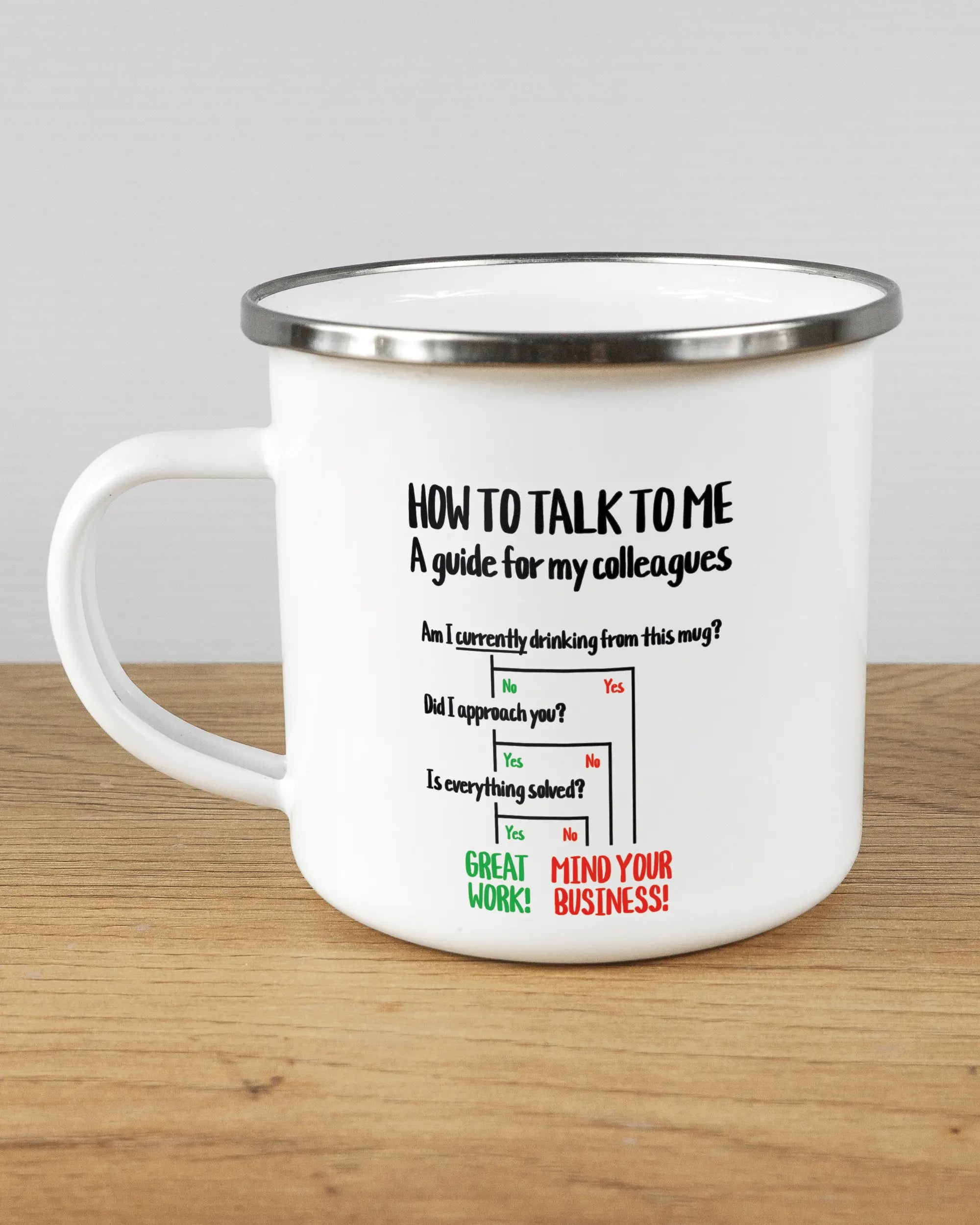 Titelbild, das die linke Seite der beidseitig bedruckten Emailletasse von Maondo mit dem Design Colleagues Guide in der Farbe White-Silver auf einem Holztisch zeigt