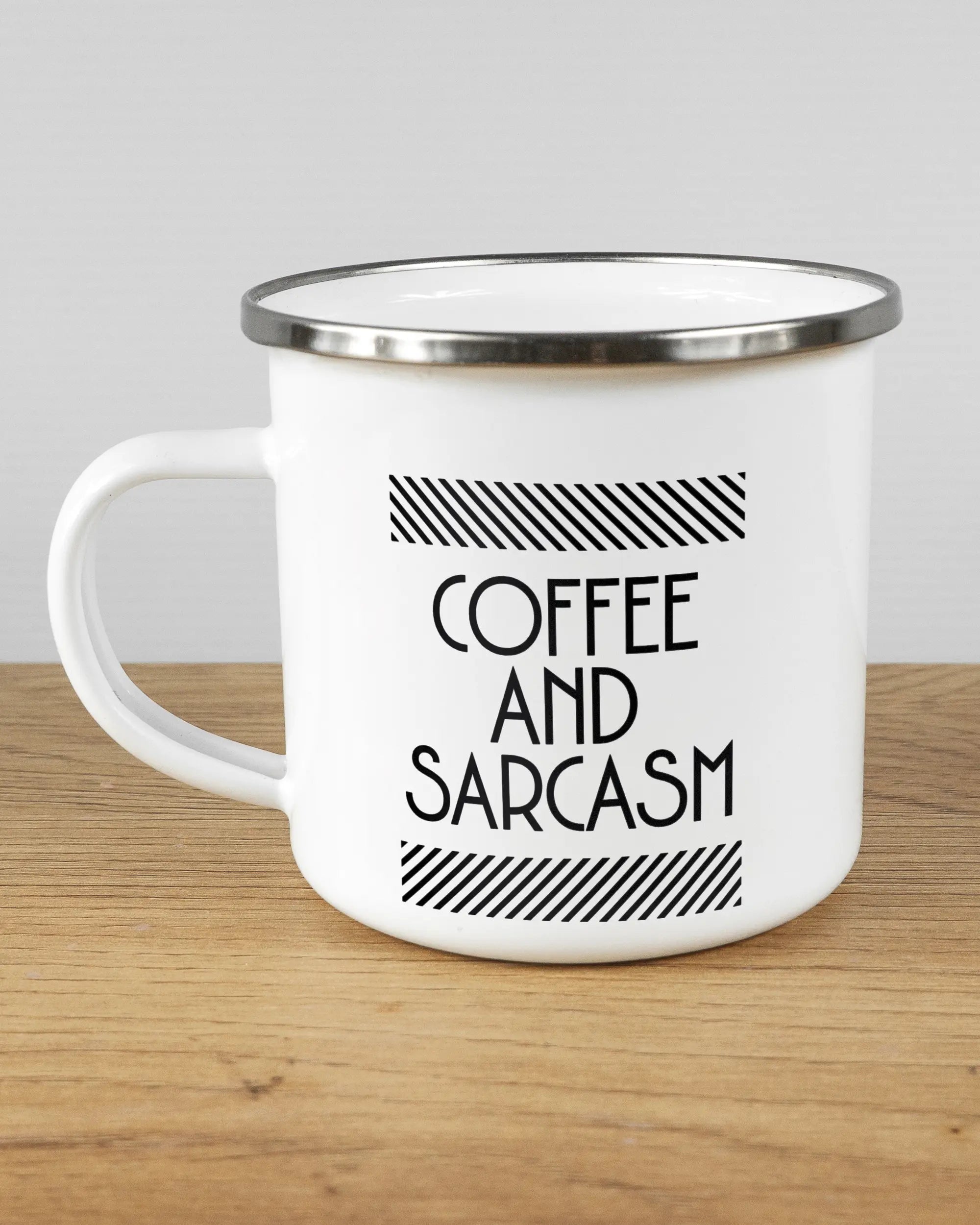 Titelbild, das die linke Seite der beidseitig bedruckten Emailletasse von Maondo mit dem Design Sarcastic Cup in der Farbe White-Silver auf einem Holztisch zeigt