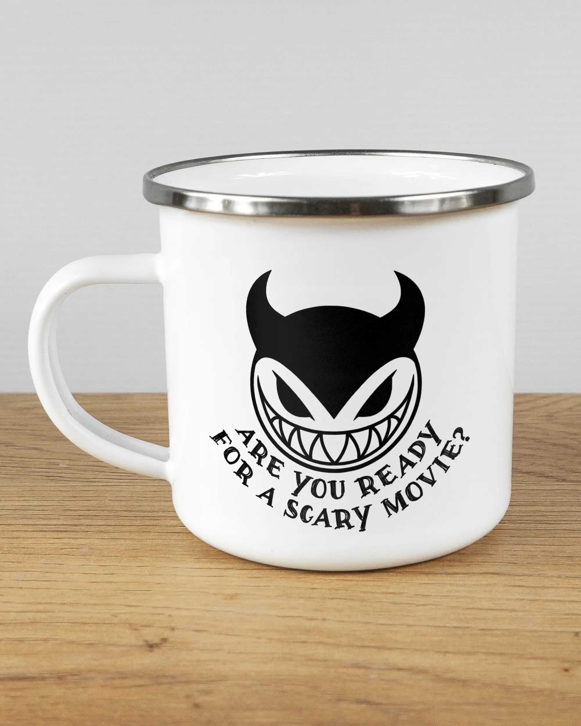 Titelbild, das die linke Seite der beidseitig bedruckten Emailletasse von Maondo mit dem Design Scary Movie in der Farbe White-Silver auf einem Holztisch zeigt