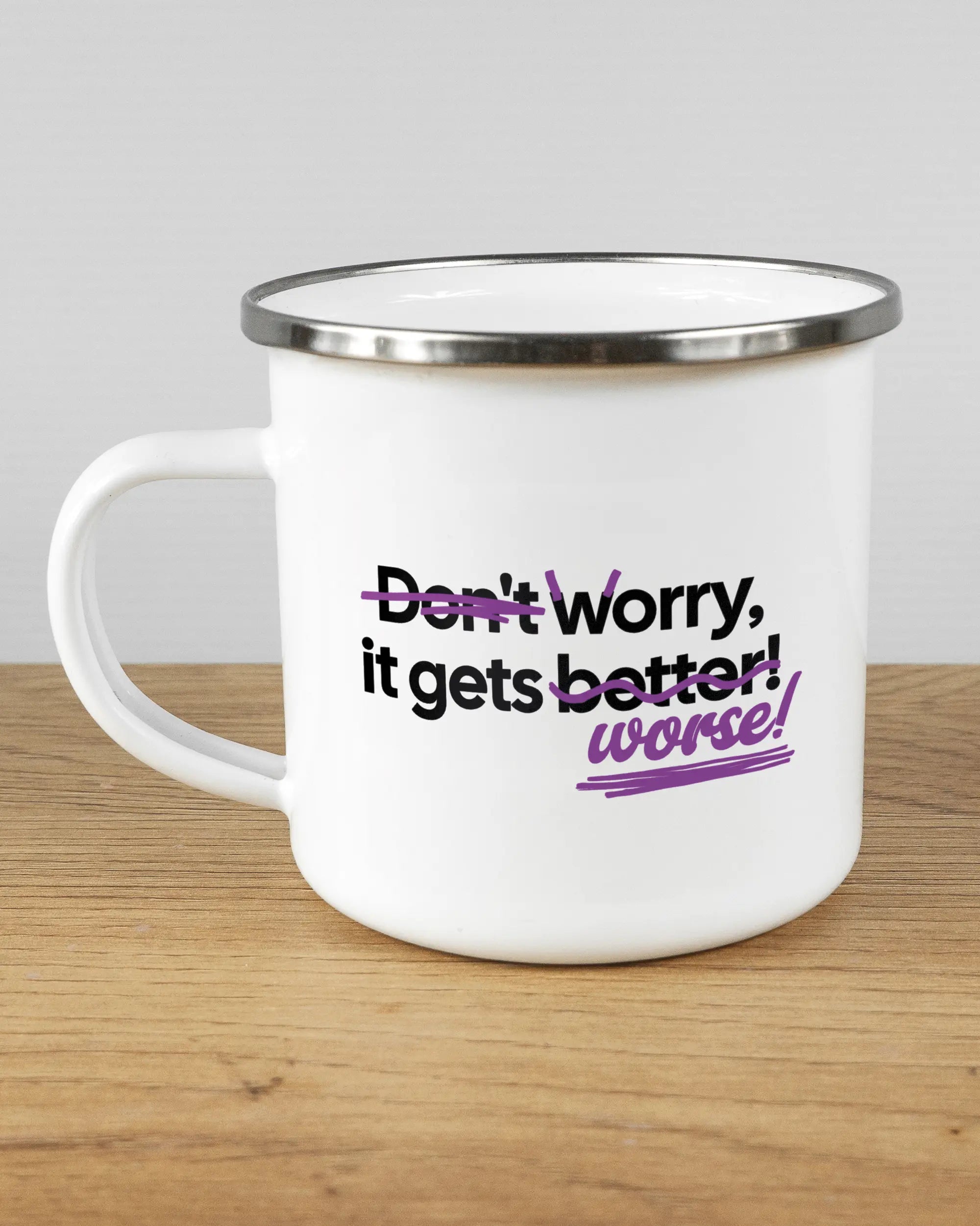 Titelbild, das die linke Seite der beidseitig bedruckten Emailletasse von Maondo mit dem Design Worry Worse in der Farbe White-Silver auf einem Holztisch zeigt