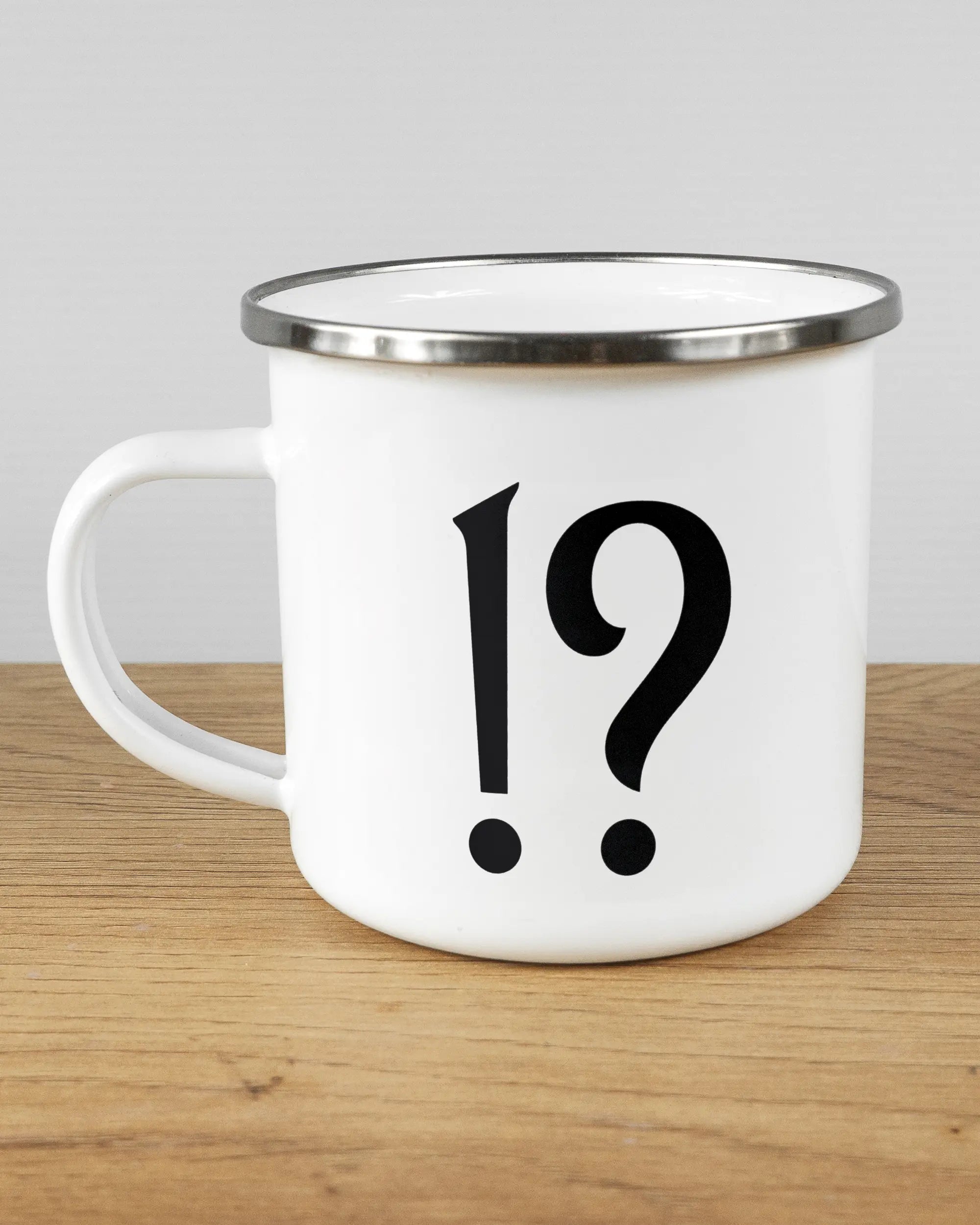 Titelbild, das die linke Seite der beidseitig bedruckten Emailletasse von Maondo mit dem Design WHAT in der Farbe White-Silver auf einem Holztisch zeigt