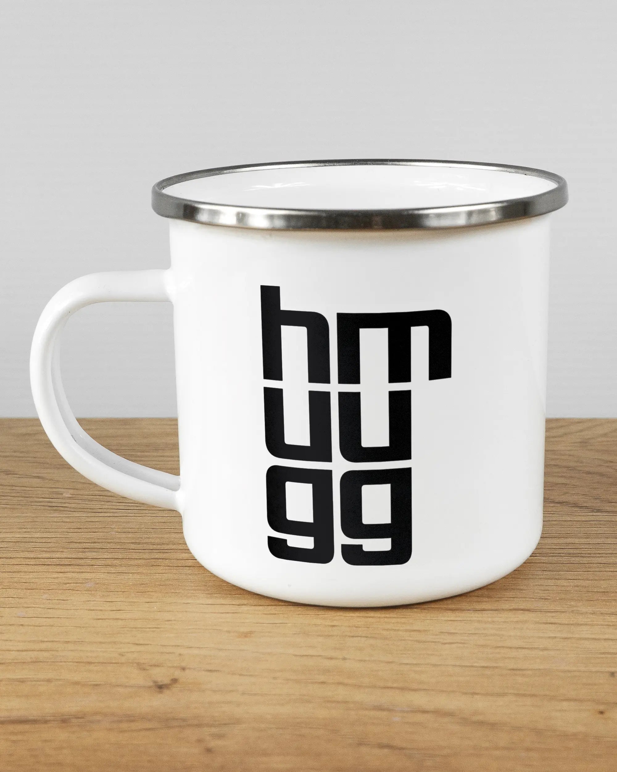 Titelbild, das die linke Seite der beidseitig bedruckten Emailletasse von Maondo mit dem Design Hug Mug in der Farbe White-Silver auf einem Holztisch zeigt