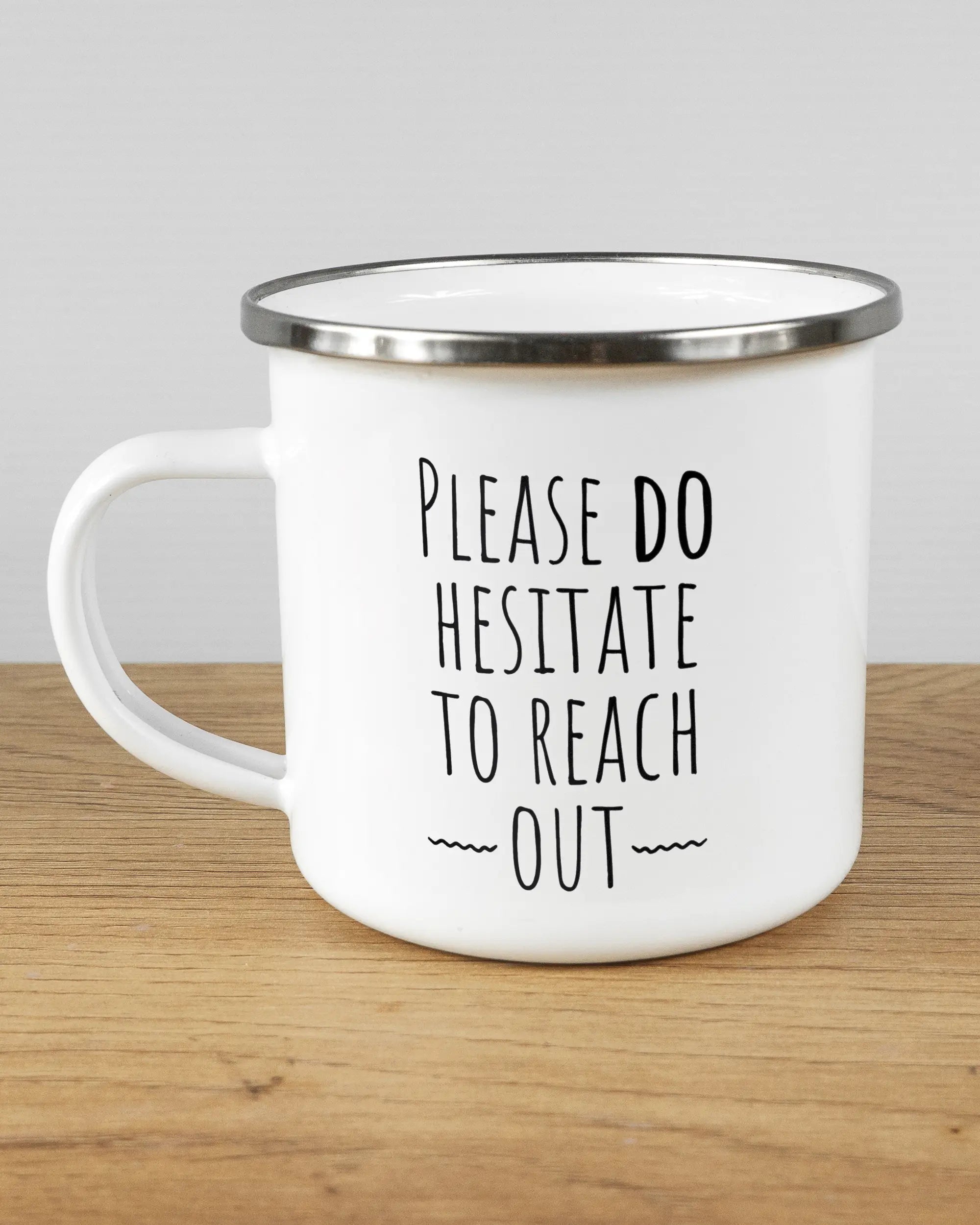 Titelbild, das die linke Seite der beidseitig bedruckten Emailletasse von Maondo mit dem Design Please Hesitate in der Farbe White-Silver auf einem Holztisch zeigt