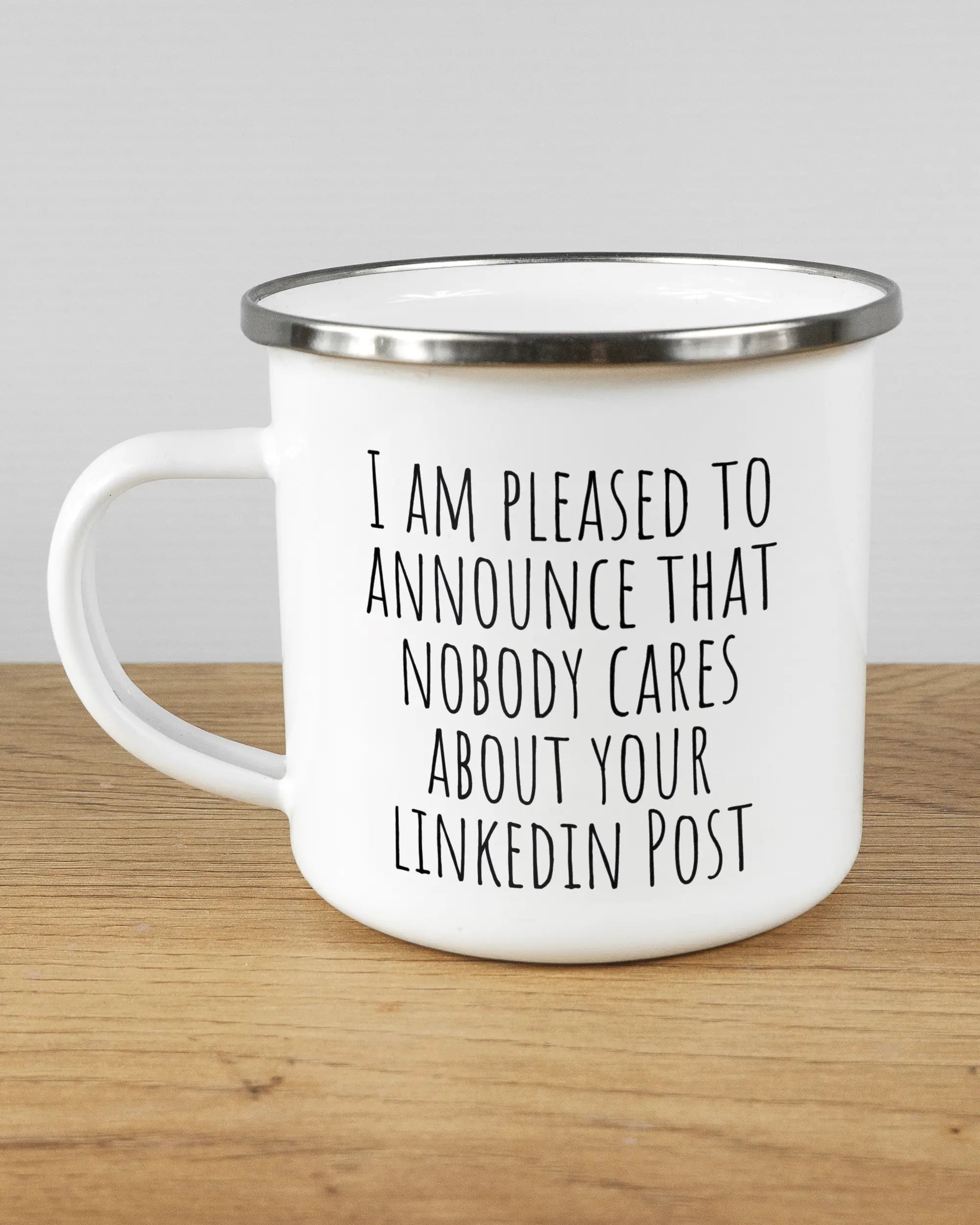 Titelbild, das die linke Seite der beidseitig bedruckten Emailletasse von Maondo mit dem Design Please Unannounce in der Farbe White-Silver auf einem Holztisch zeigt