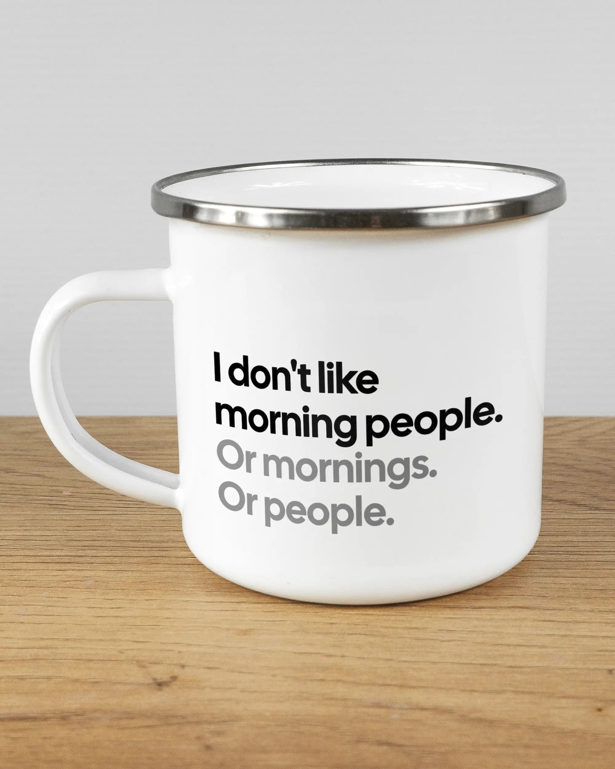 Titelbild, das die linke Seite der beidseitig bedruckten Emailletasse von Maondo mit dem Design Morning Person in der Farbe White-Silver auf einem Holztisch zeigt