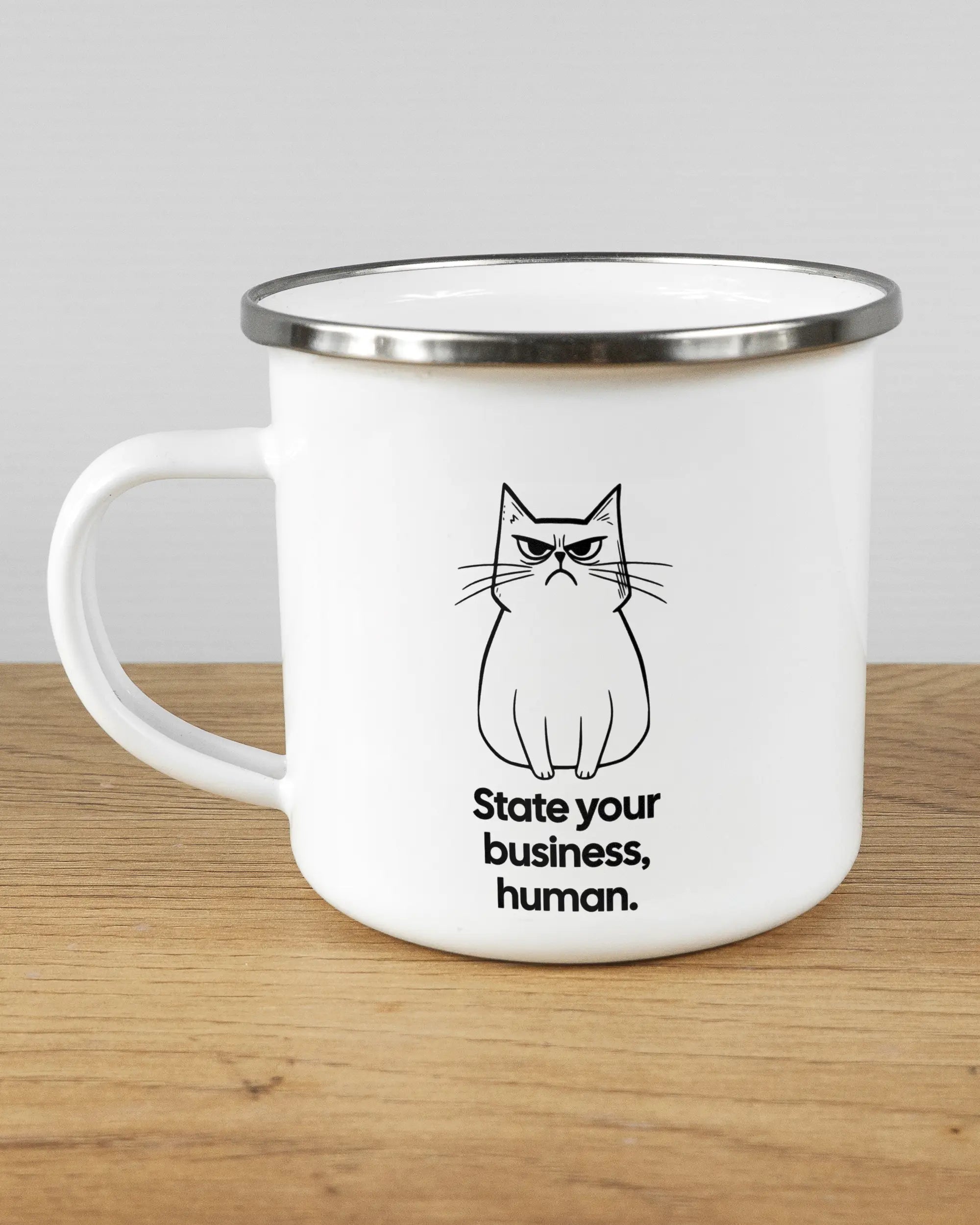 Titelbild, das die linke Seite der beidseitig bedruckten Emailletasse von Maondo mit dem Design Grumpy Purr in der Farbe White-Silver auf einem Holztisch zeigt