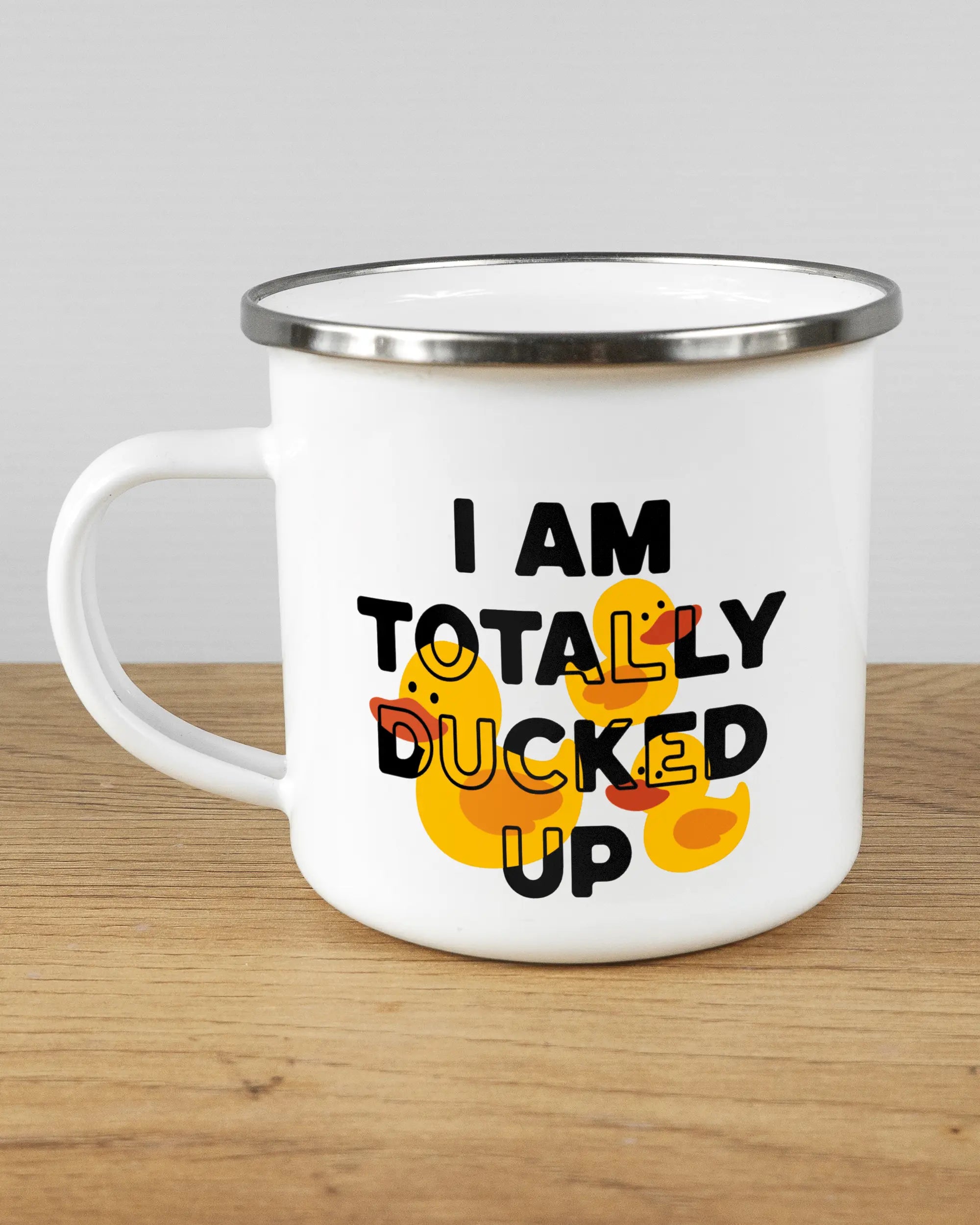 Titelbild, das die linke Seite der beidseitig bedruckten Emailletasse von Maondo mit dem Design Ducked Up in der Farbe White-Silver auf einem Holztisch zeigt