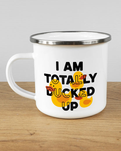 Titelbild, das die linke Seite der beidseitig bedruckten Emailletasse von Maondo mit dem Design Ducked Up in der Farbe White-Silver auf einem Holztisch zeigt