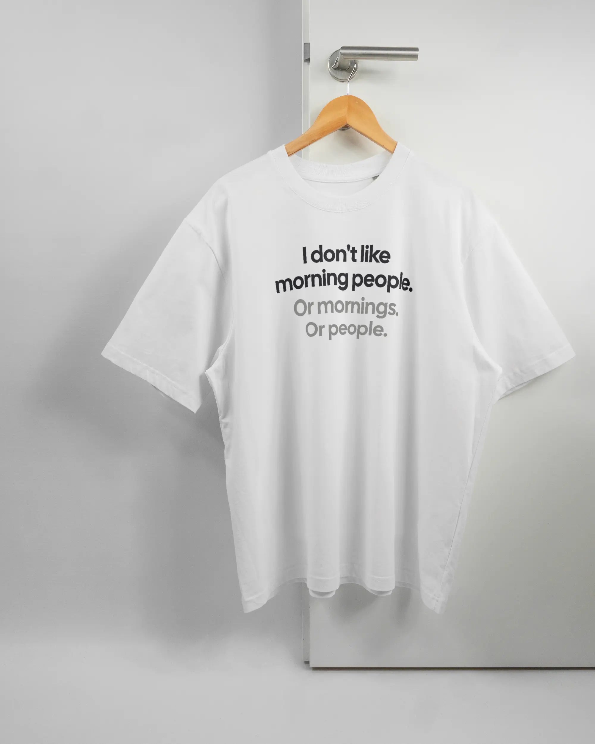 Vorderseite des Basic Unisex T-Shirt Oversized von Maondo mit dem Design Morning Person in der Farbe White und in der Größe L auf einem Bügel an der Türklinke aufgehängt