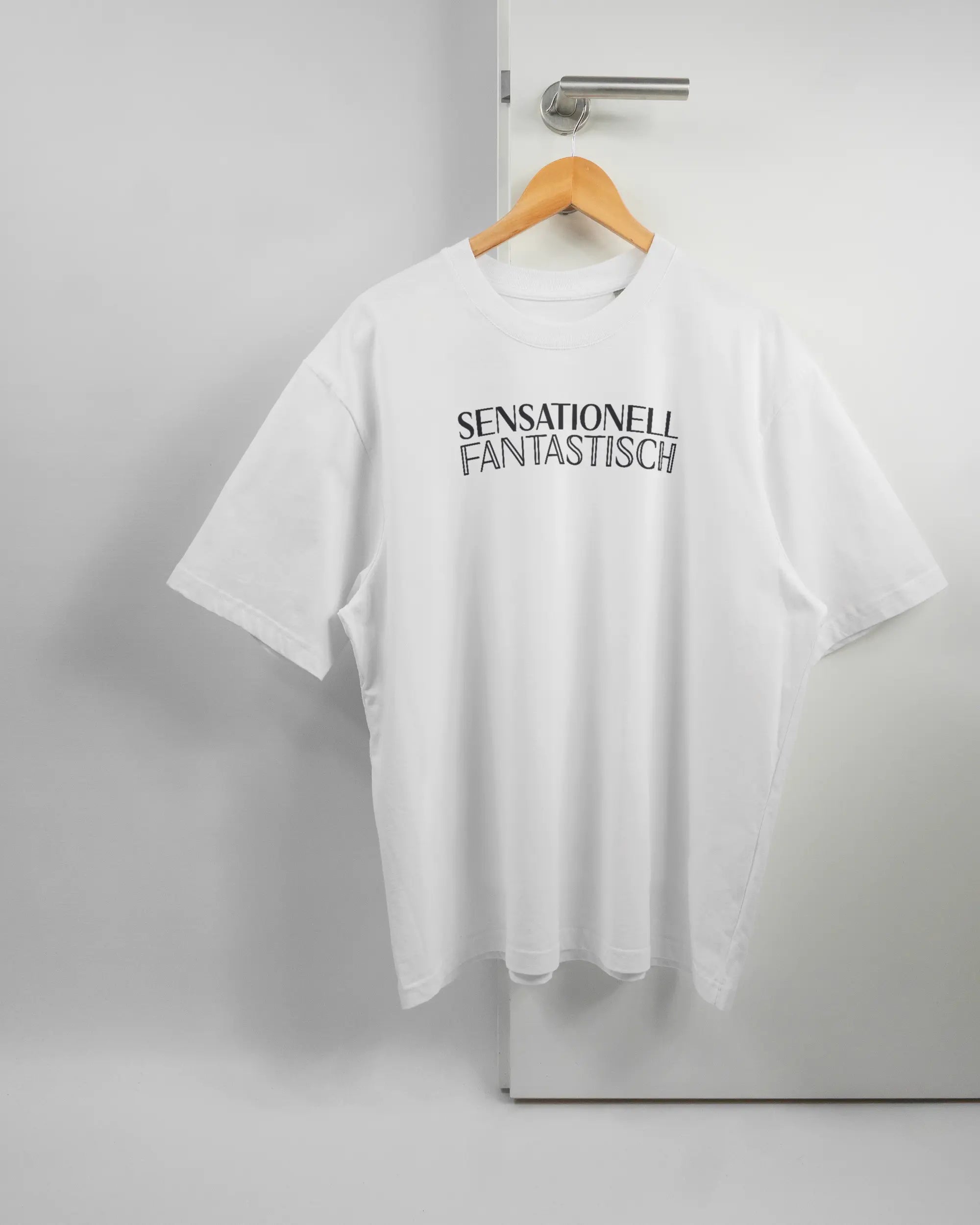 Vorderseite des Basic Unisex T-Shirt Oversized von Maondo mit dem Design Sensationell in der Farbe White und in der Größe L auf einem Bügel an der Türklinke aufgehängt