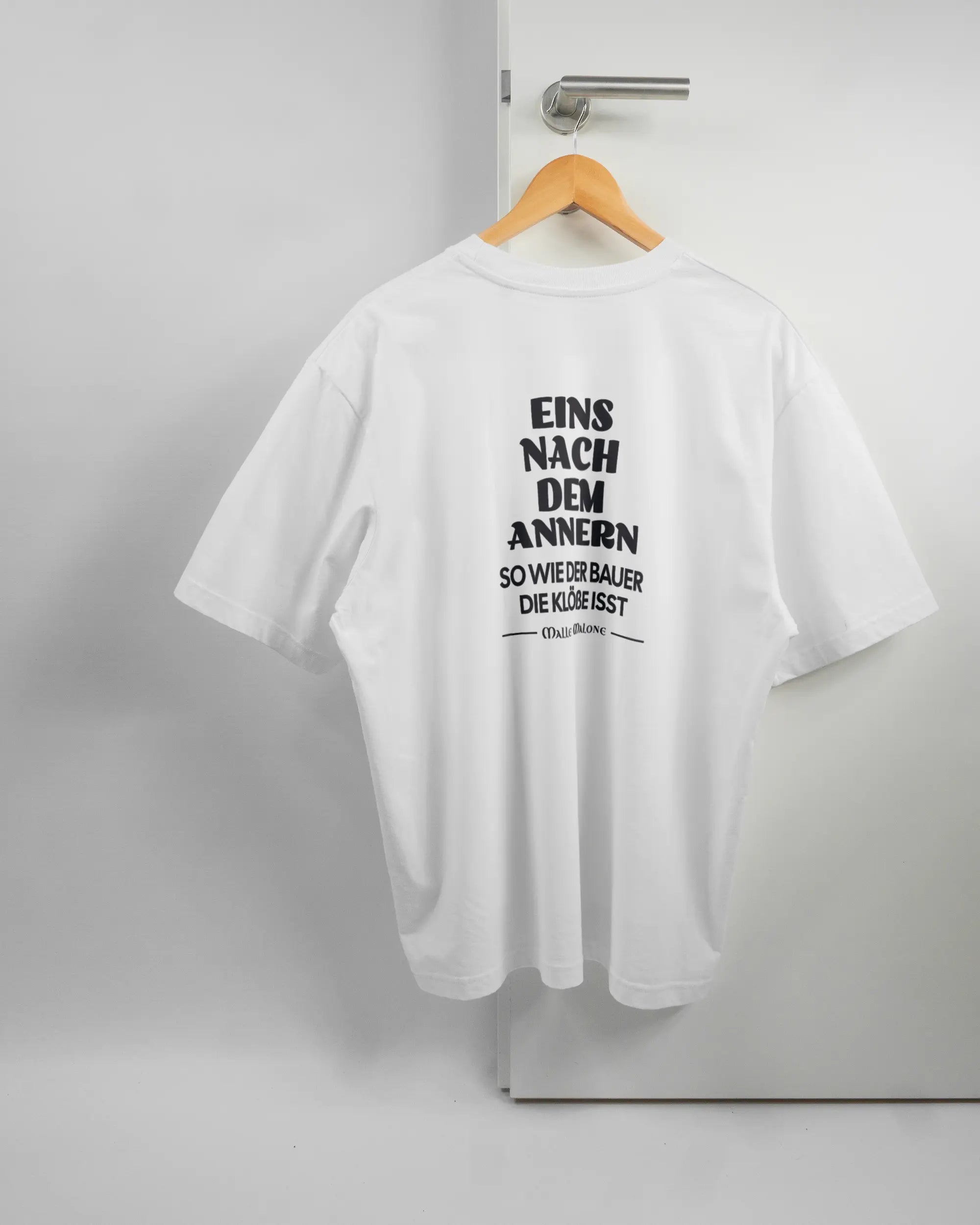 Rückseite des Basic Unisex T-Shirt Oversized von Maondo mit dem Design Eins nach dem Annern in der Farbe White und in der Größe L auf einem Bügel an der Türklinke aufgehängt