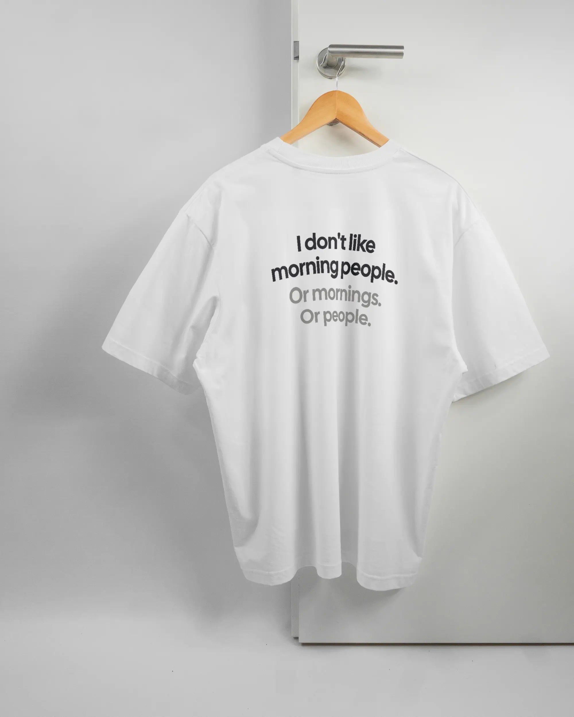 Rückseite des Basic Unisex T-Shirt Oversized von Maondo mit dem Design Morning Person in der Farbe White und in der Größe L auf einem Bügel an der Türklinke aufgehängt