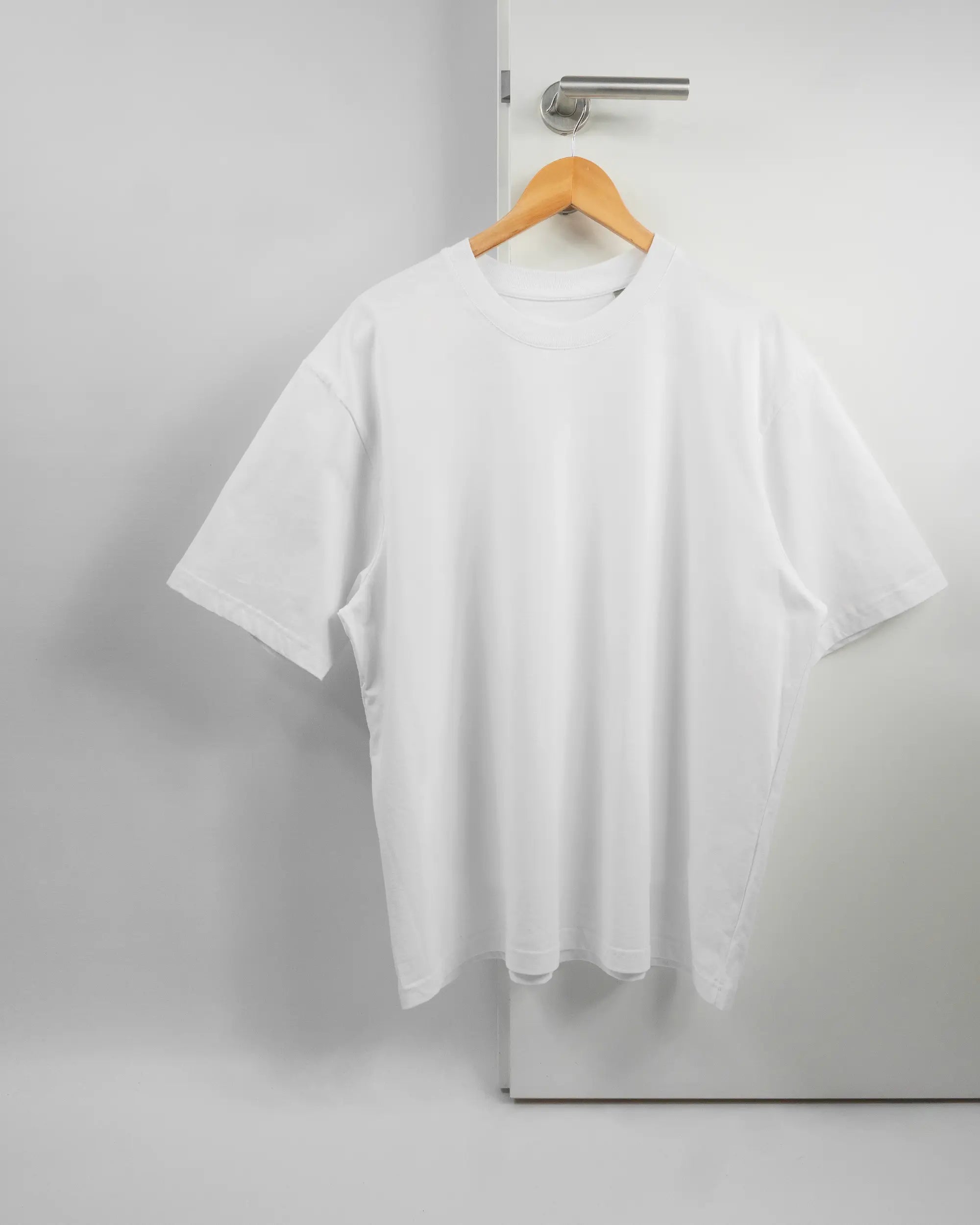 Vorderseite des Basic Unisex T-Shirt Oversized von Maondo ohne Motiv in der Farbe White und in der Größe L auf einem Bügel an der Türklinke aufgehängt