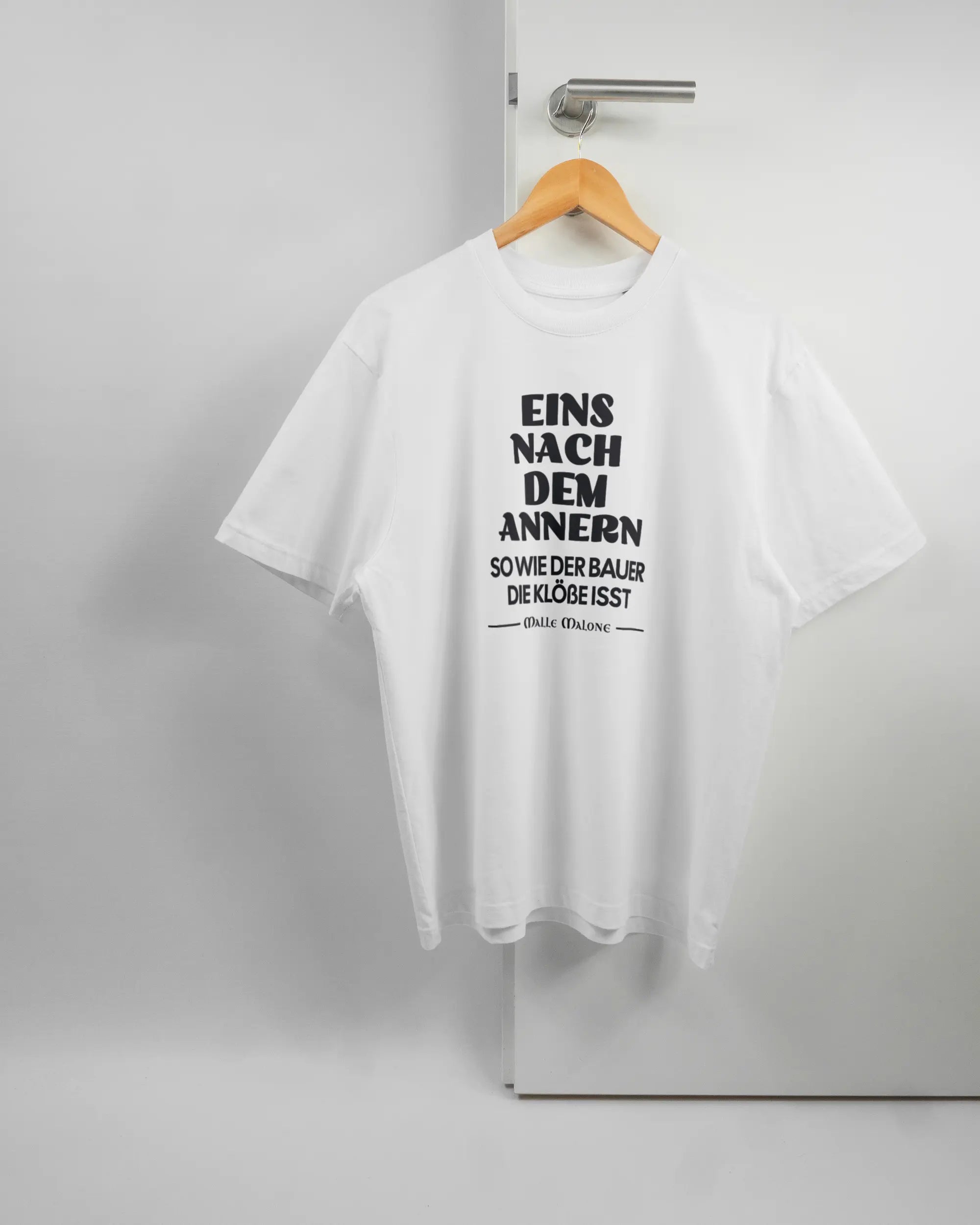 Vorderseite des Basic Unisex T-Shirt Oversized von Maondo mit dem Design Eins nach dem Annern in der Farbe White und in der Größe S auf einem Bügel an der Türklinke aufgehängt