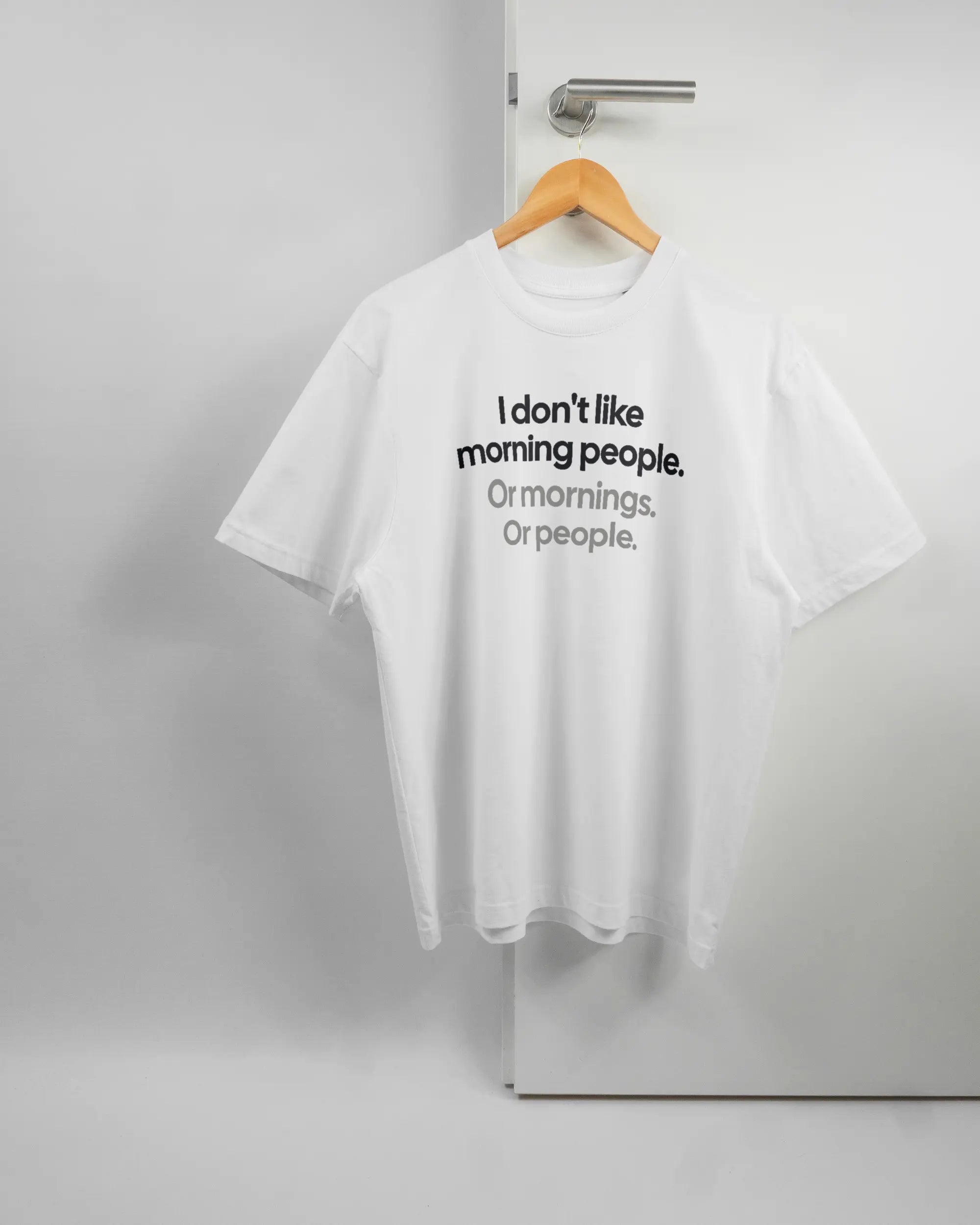 Vorderseite des Basic Unisex T-Shirt Oversized von Maondo mit dem Design Morning Person in der Farbe White und in der Größe S auf einem Bügel an der Türklinke aufgehängt