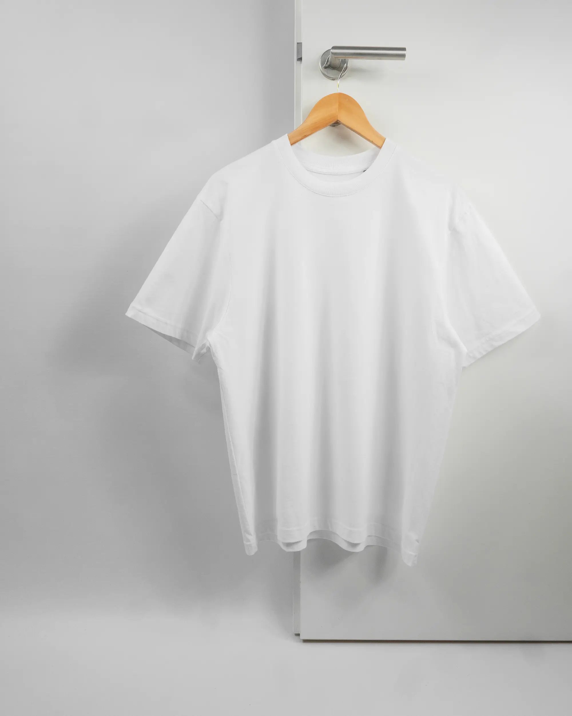 Vorderseite des Basic Unisex T-Shirt Oversized von Maondo ohne Motiv in der Farbe White und in der Größe S auf einem Bügel an der Türklinke aufgehängt