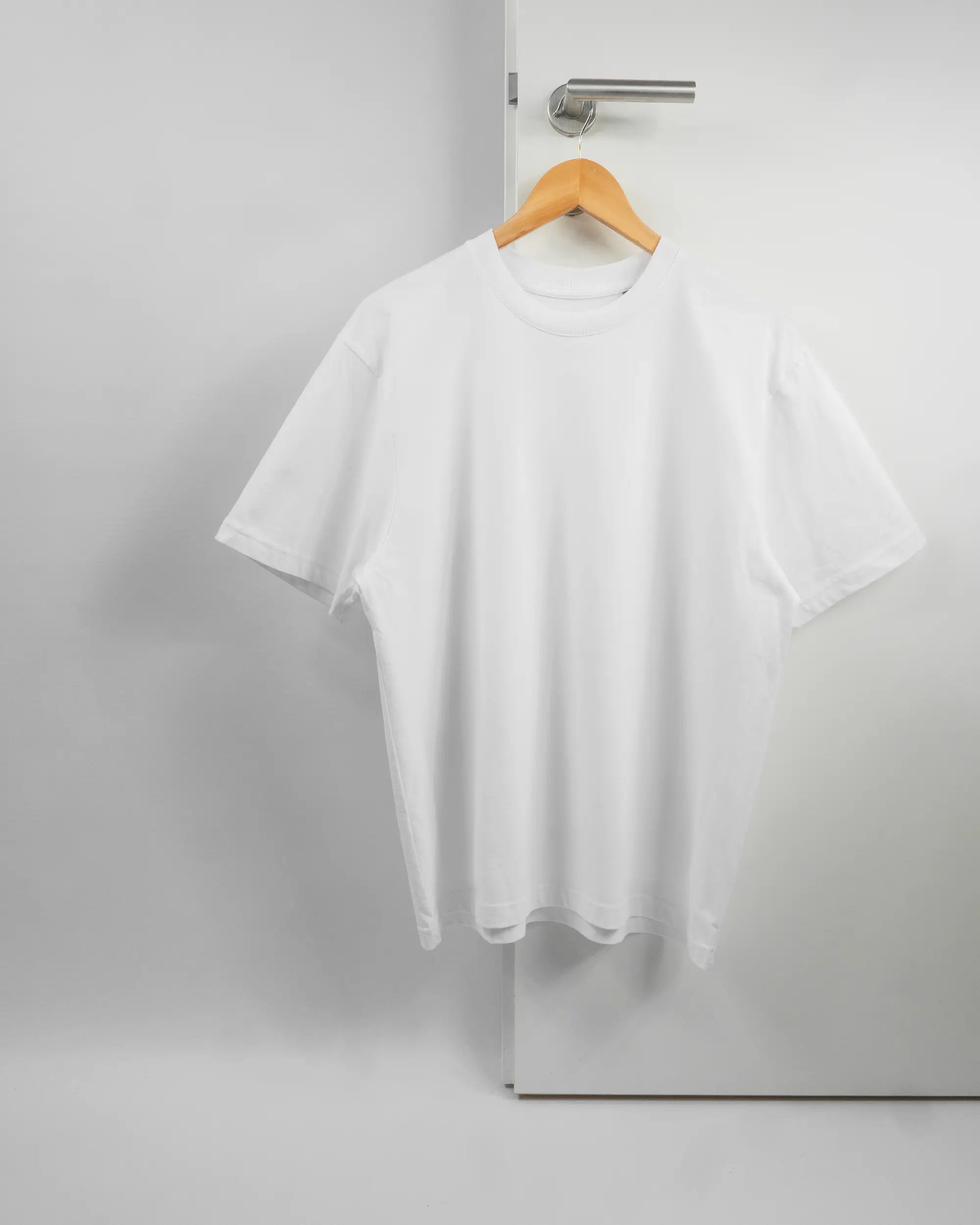 Vorderseite des Basic Unisex T-Shirt Oversized von Maondo ohne Motiv in der Farbe White und in der Größe S auf einem Bügel an der Türklinke aufgehängt