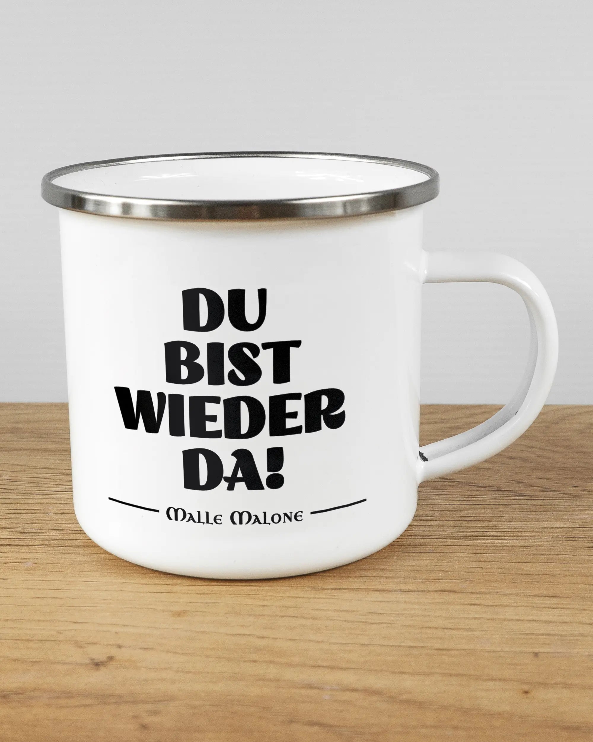 Alternativbild, das die rechte Seite der beidseitig bedruckten Emailletasse von Maondo mit dem Design Wieder da in der Farbe White-Silver auf einem Holztisch zeigt