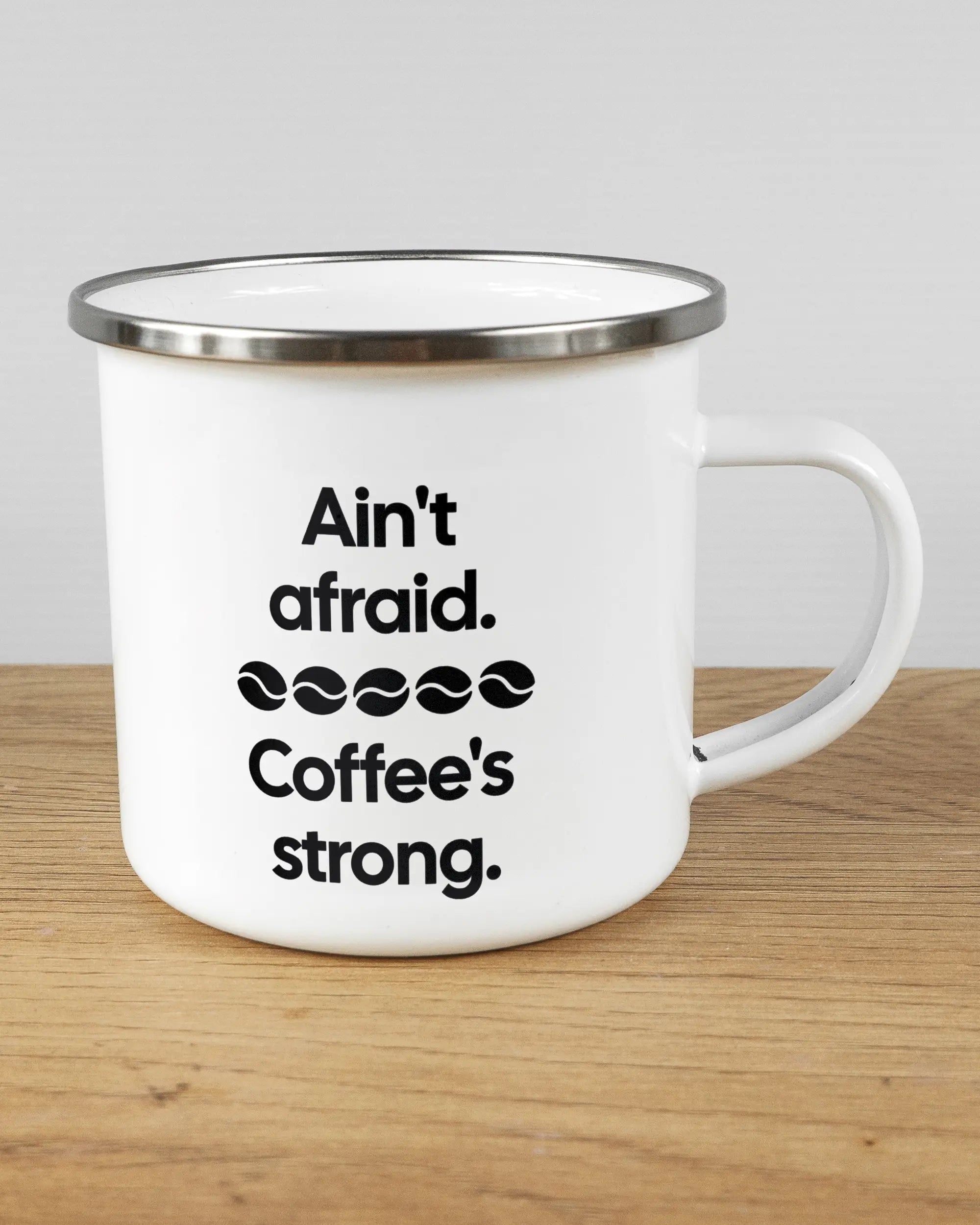 Alternativbild, das die rechte Seite der beidseitig bedruckten Emailletasse von Maondo mit dem Design Coffee is strong in der Farbe White-Silver auf einem Holztisch zeigt