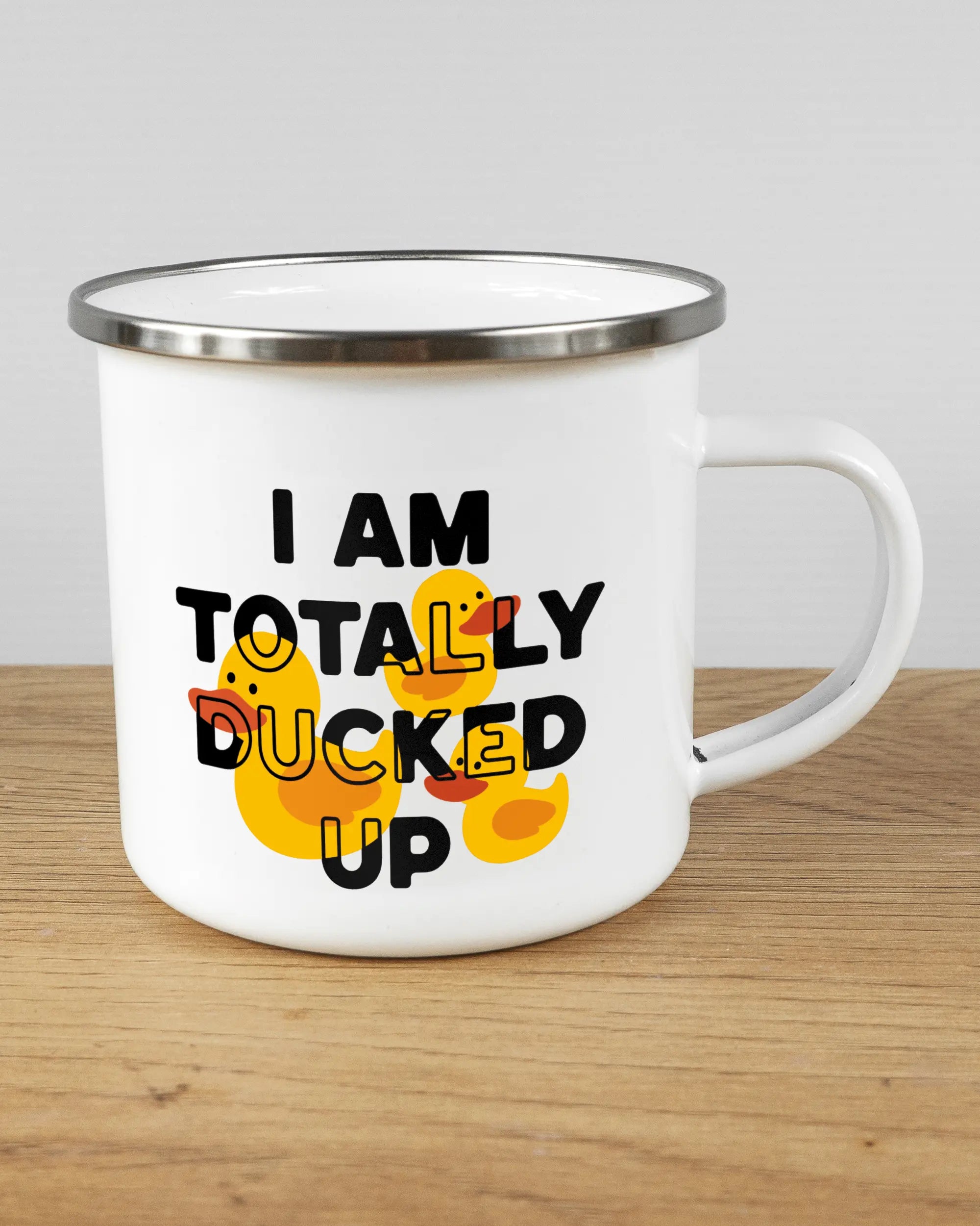 Alternativbild, das die rechte Seite der beidseitig bedruckten Emailletasse von Maondo mit dem Design Ducked Up in der Farbe White-Silver auf einem Holztisch zeigt