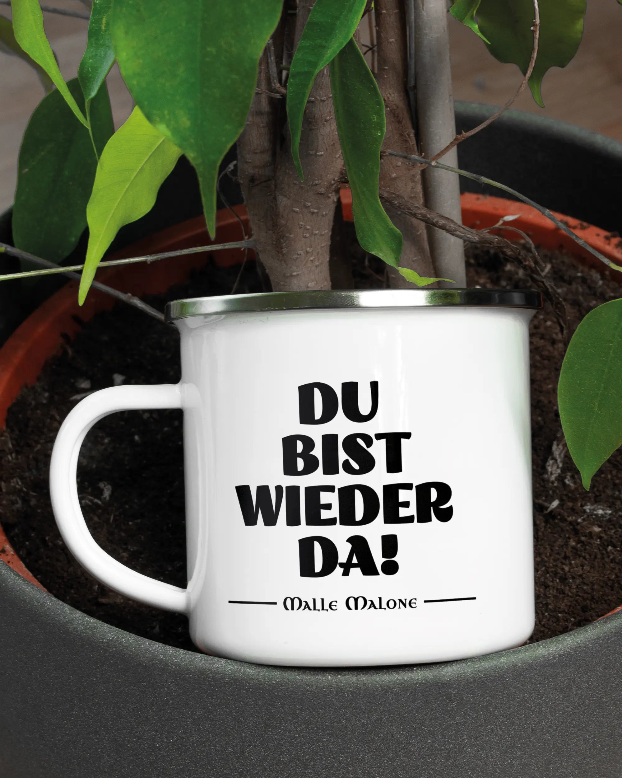Aufnahme der linken Seite der beidseitig bedruckten Emailletasse von Maondo mit dem Design Wieder da in der Farbe White-Silver in einem Blumentopf