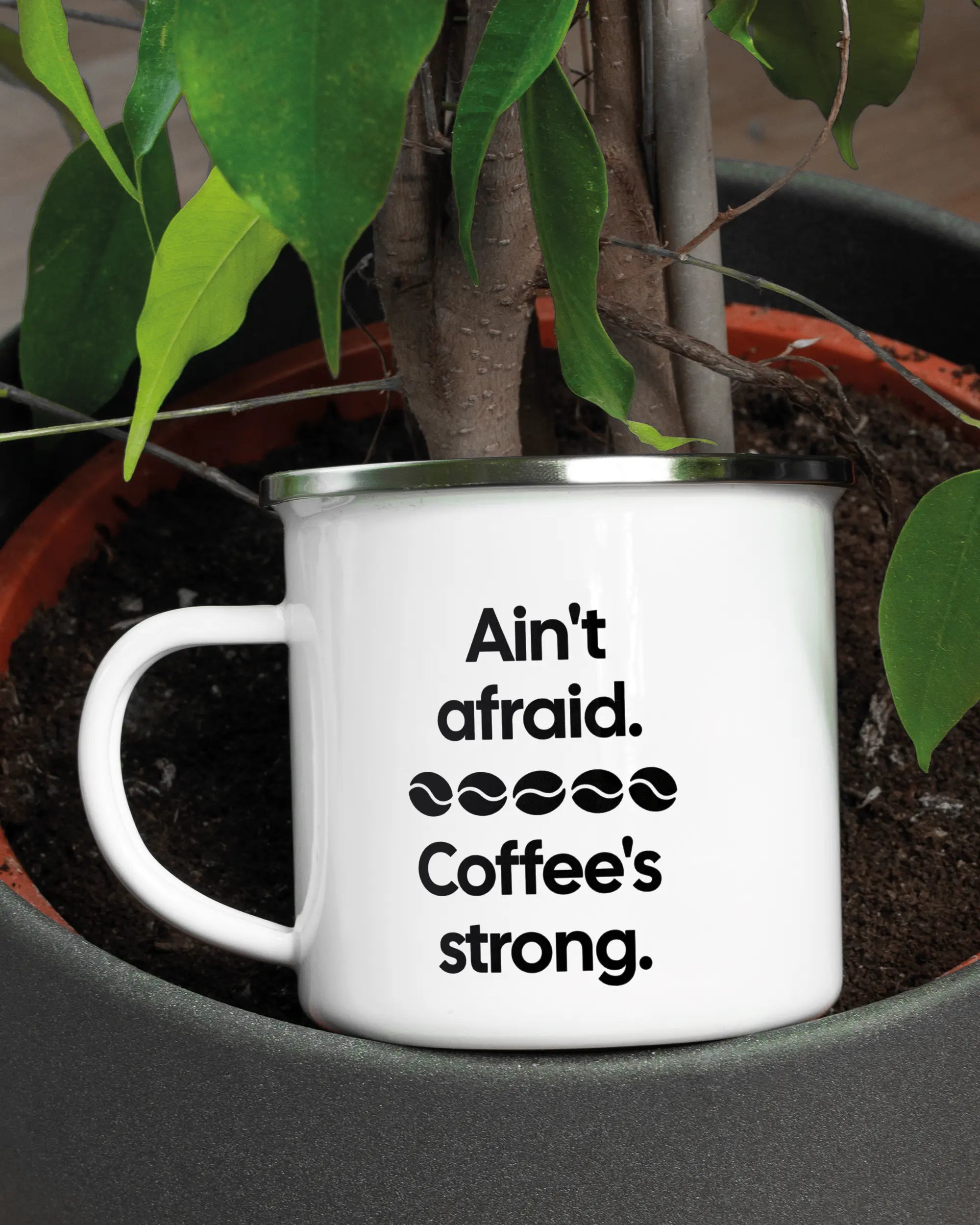 Aufnahme der linken Seite der beidseitig bedruckten Emailletasse von Maondo mit dem Design Coffee is strong in der Farbe White-Silver in einem Blumentopf