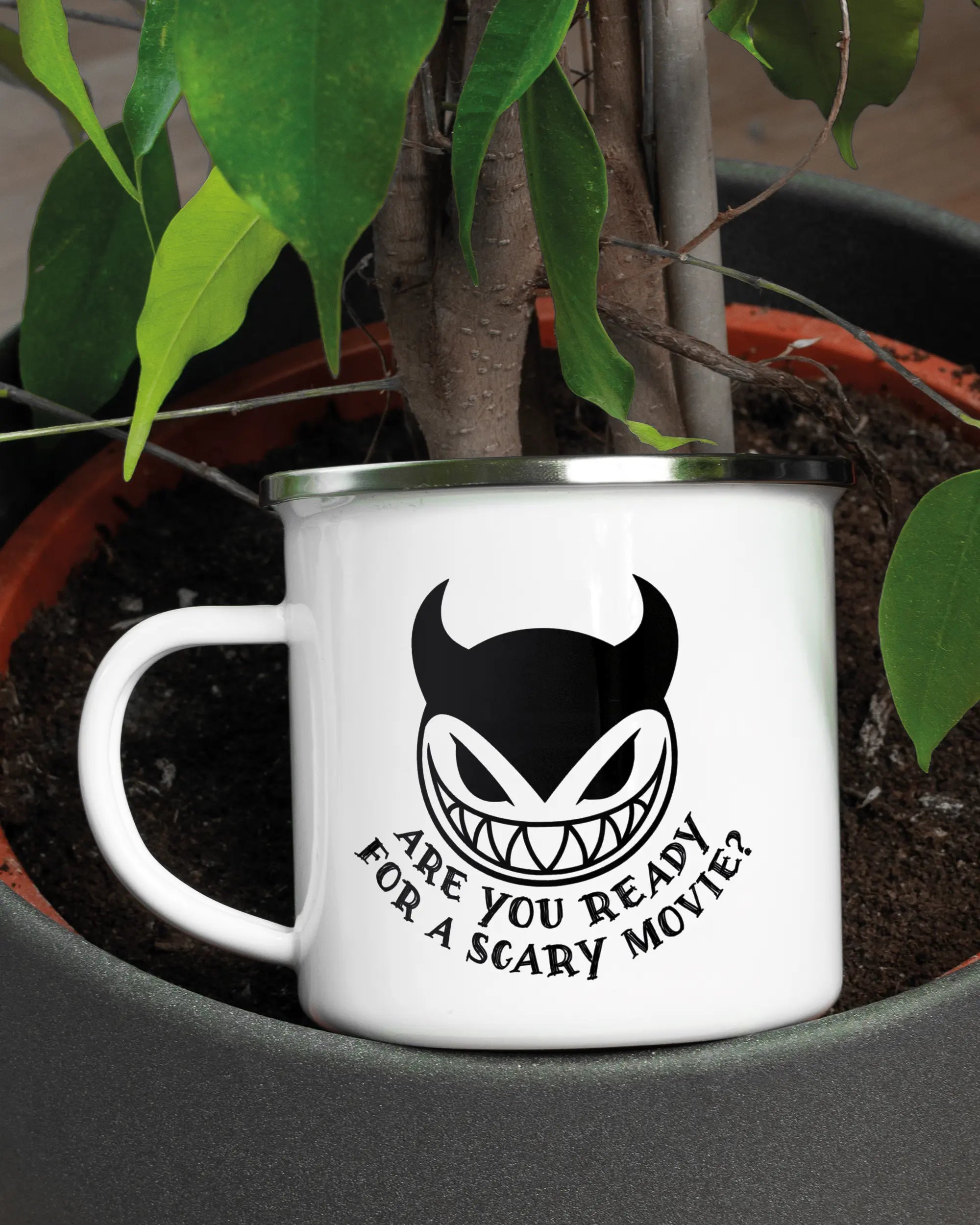 Aufnahme der linken Seite der beidseitig bedruckten Emailletasse von Maondo mit dem Design Scary Movie in der Farbe White-Silver in einem Blumentopf