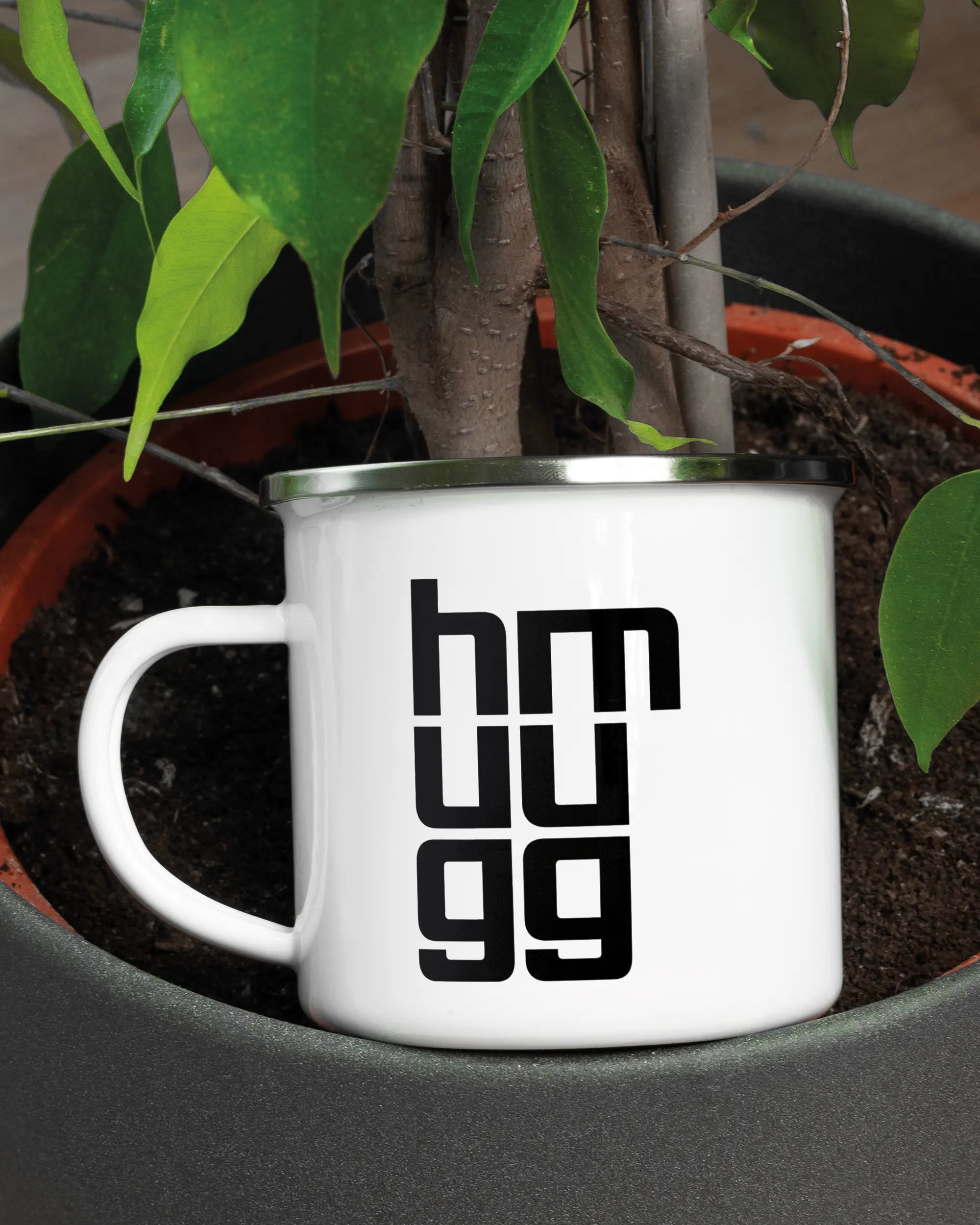 Aufnahme der linken Seite der beidseitig bedruckten Emailletasse von Maondo mit dem Design Hug Mug in der Farbe White-Silver in einem Blumentopf
