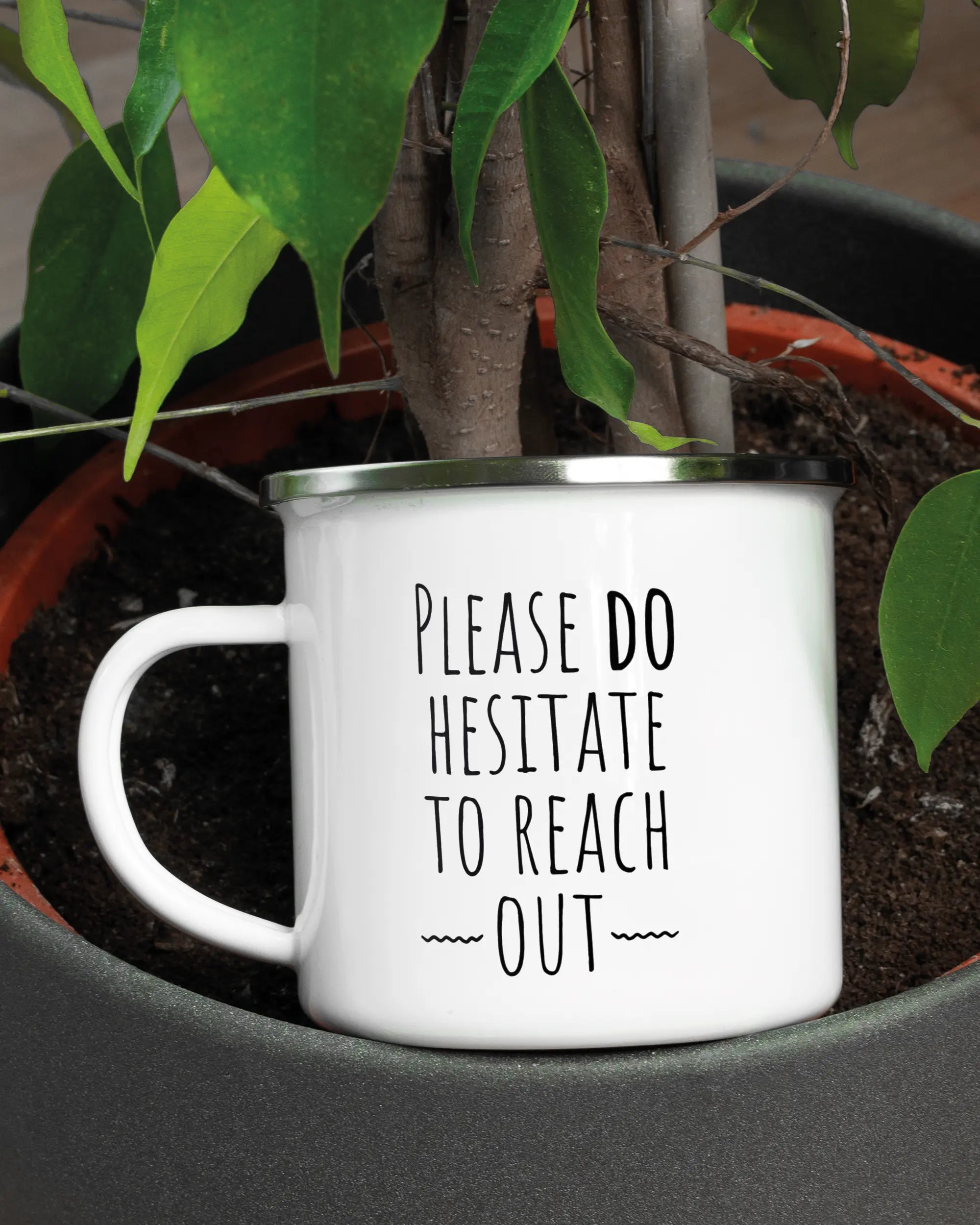 Aufnahme der linken Seite der beidseitig bedruckten Emailletasse von Maondo mit dem Design Please Hesitate in der Farbe White-Silver in einem Blumentopf