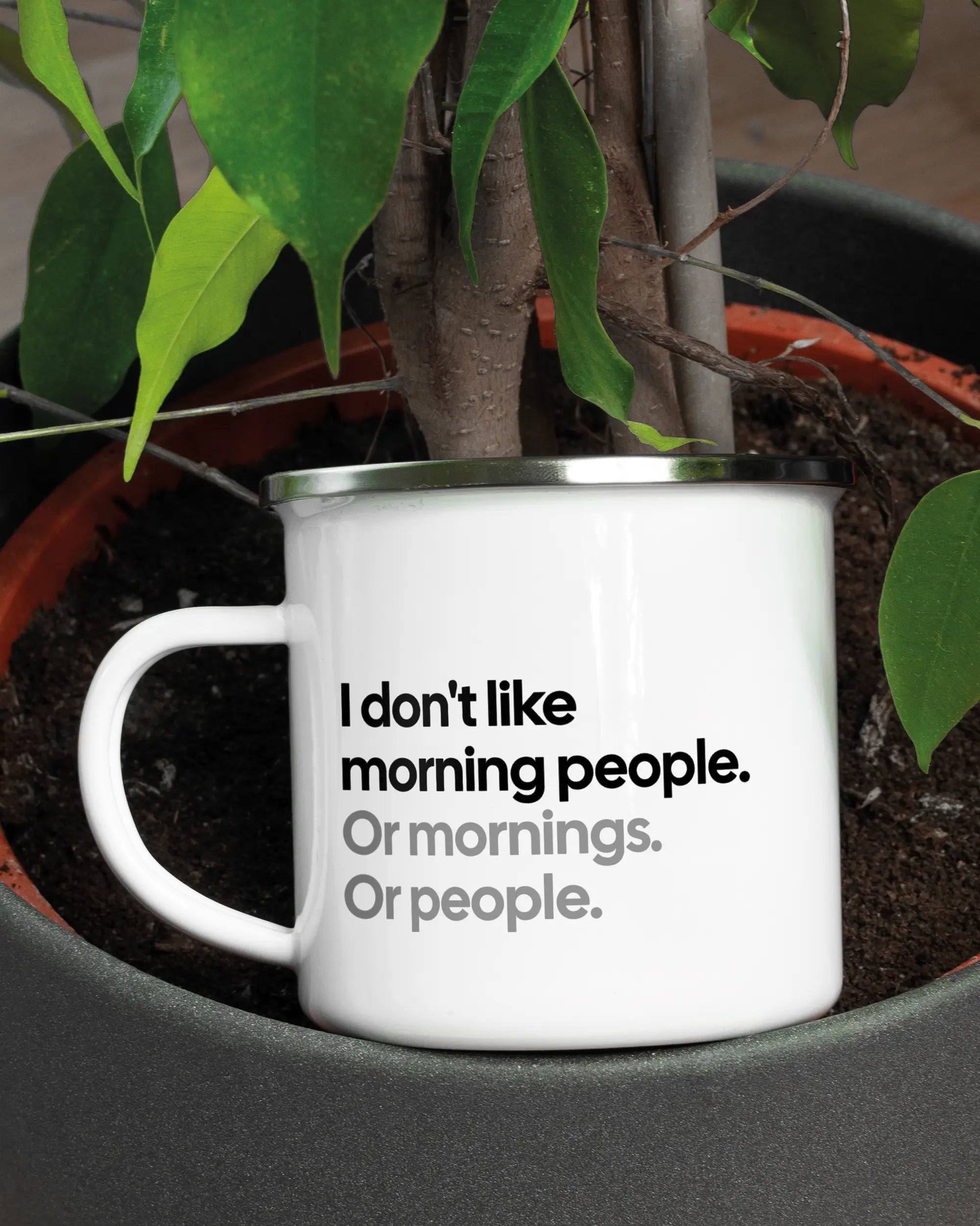 Aufnahme der linken Seite der beidseitig bedruckten Emailletasse von Maondo mit dem Design Morning Person in der Farbe White-Silver in einem Blumentopf