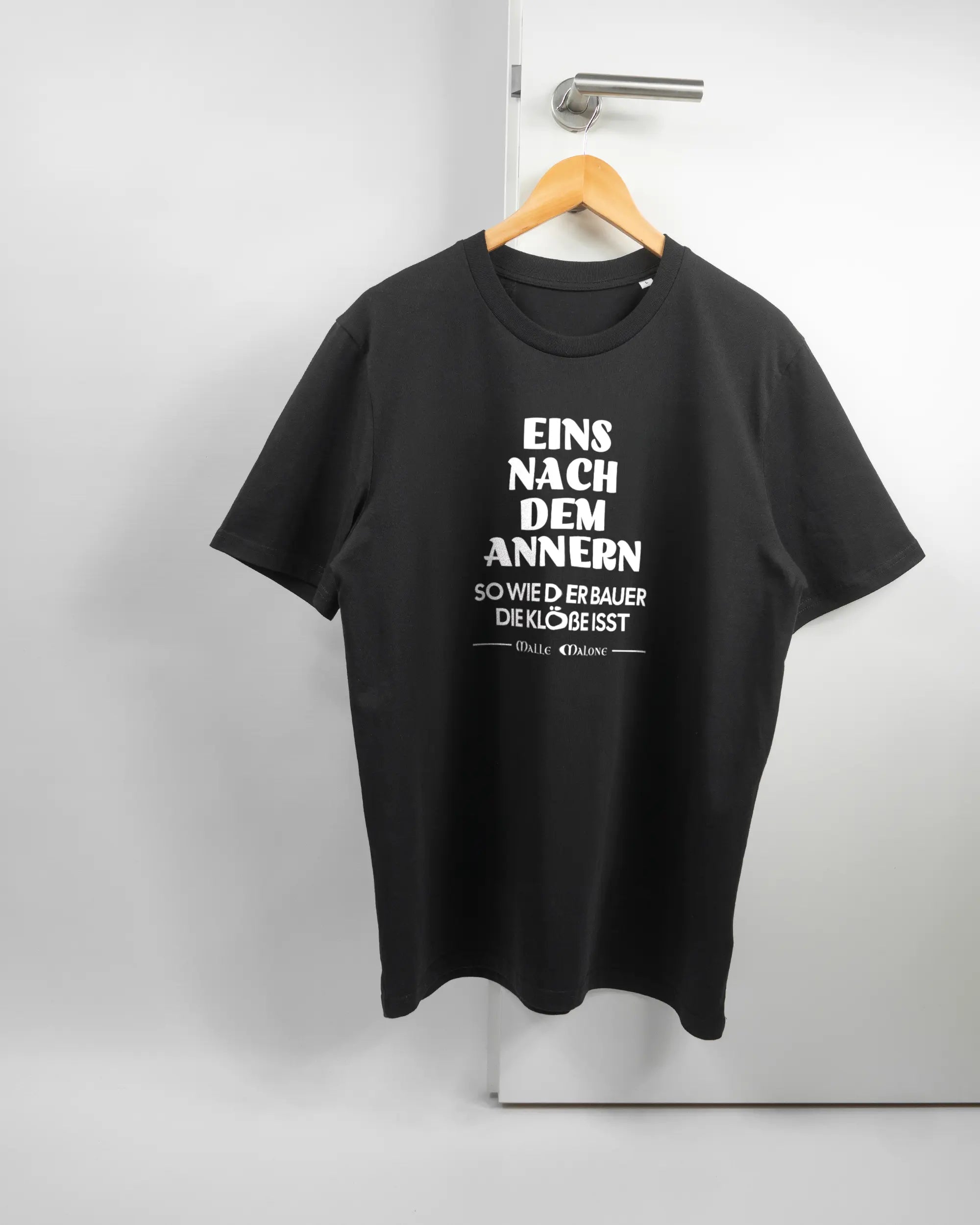Vorderseite des Organic Unisex T-Shirt Casual von Maondo mit dem Design Eins nach dem Annern in der Farbe Black und in der Größe L auf einem Bügel an der Türklinke aufgehängt