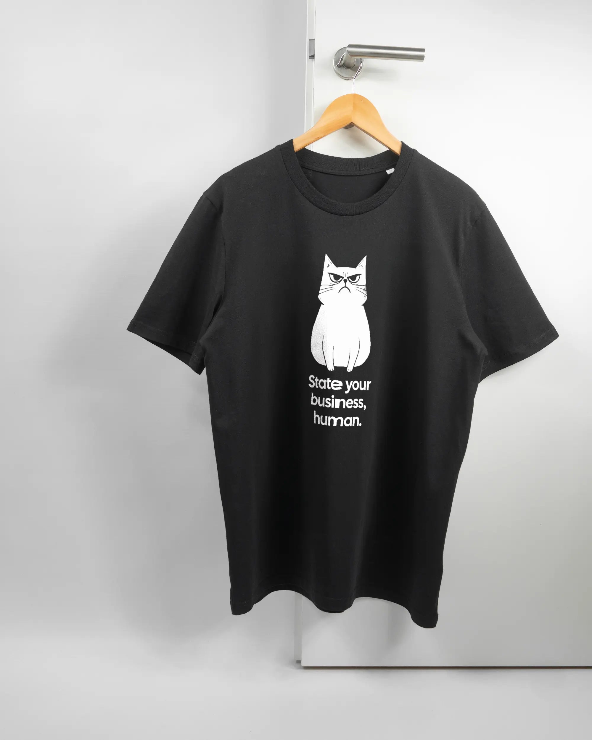 Vorderseite des Organic Unisex T-Shirt Casual von Maondo mit dem Design Grumpy Purr in der Farbe Black und in der Größe L auf einem Bügel an der Türklinke aufgehängt