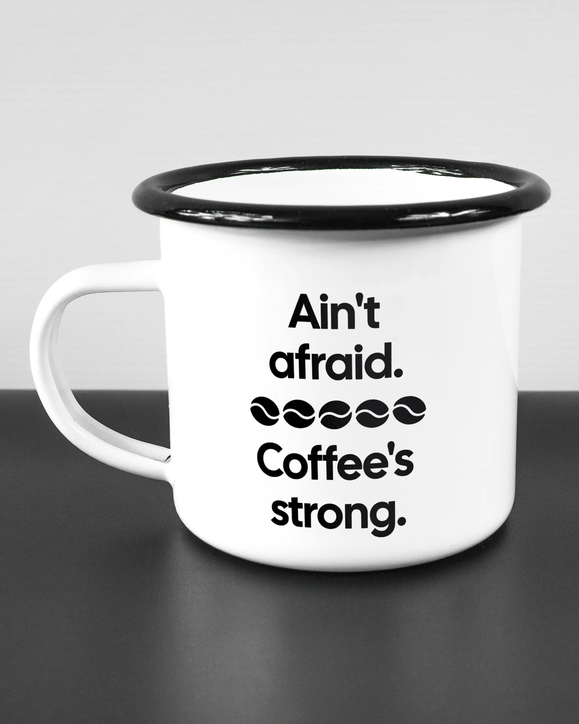 Titelbild, das die linke Seite der beidseitig bedruckten Emailletasse von Maondo mit dem Design Coffee is strong in der Farbe White-Black auf einem Holztisch zeigt