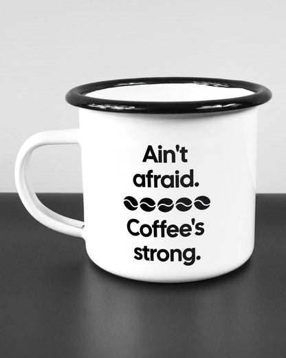 Titelbild, das die linke Seite der beidseitig bedruckten Emailletasse von Maondo mit dem Design Coffee is strong in der Farbe White-Black auf einem Holztisch zeigt