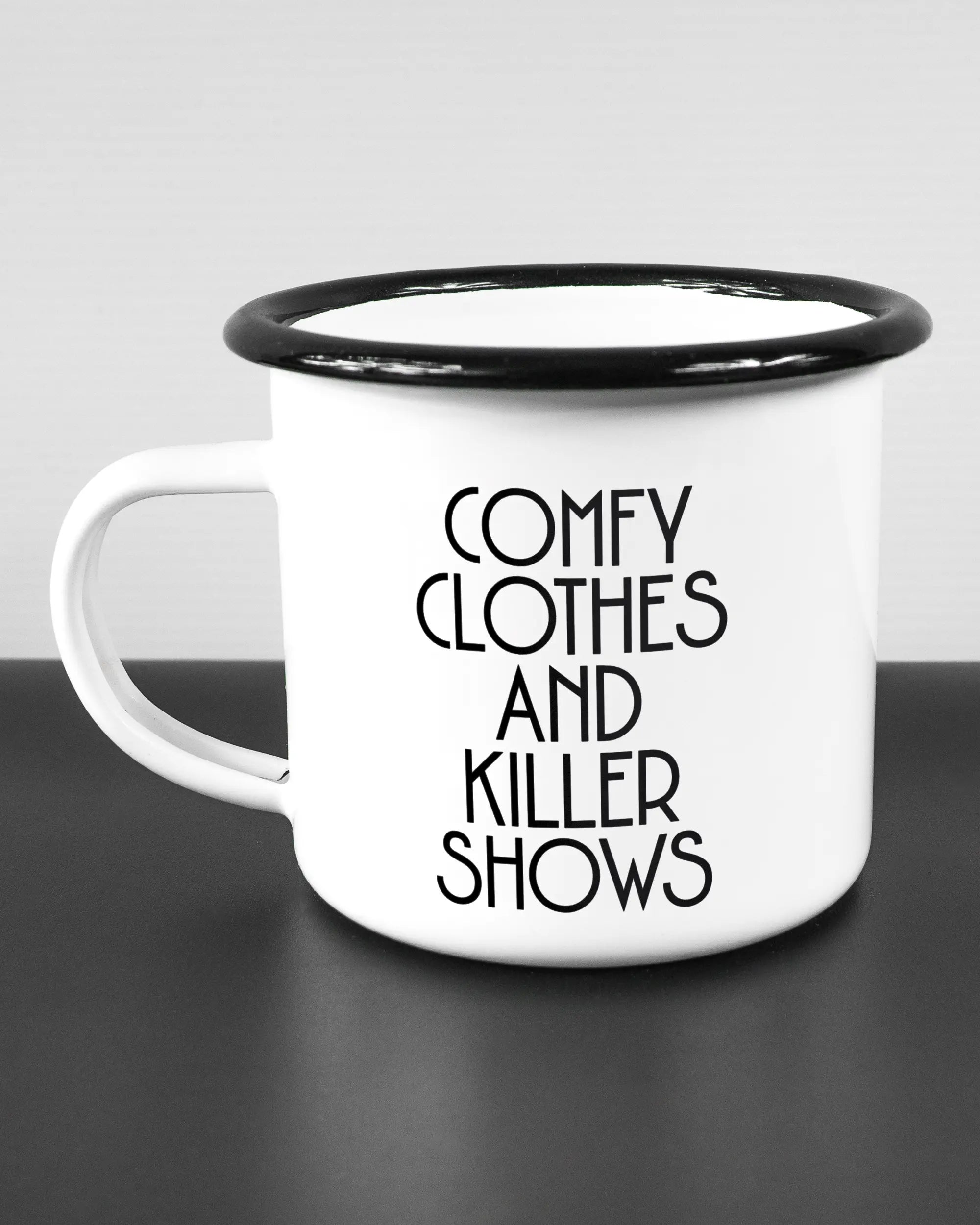 Titelbild, das die linke Seite der beidseitig bedruckten Emailletasse von Maondo mit dem Design Killer Shows in der Farbe White-Black auf einem Holztisch zeigt