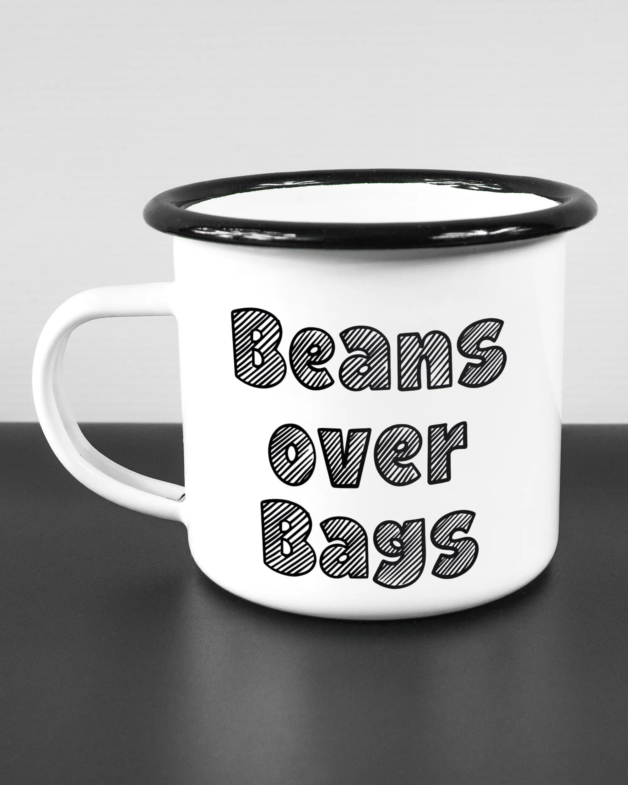 Titelbild, das die linke Seite der beidseitig bedruckten Emailletasse von Maondo mit dem Design Beans over Bags in der Farbe White-Black auf einem Holztisch zeigt