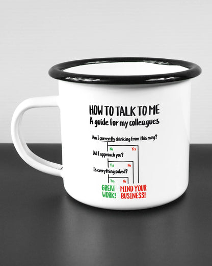 Titelbild, das die linke Seite der beidseitig bedruckten Emailletasse von Maondo mit dem Design Colleagues Guide in der Farbe White-Black auf einem Holztisch zeigt