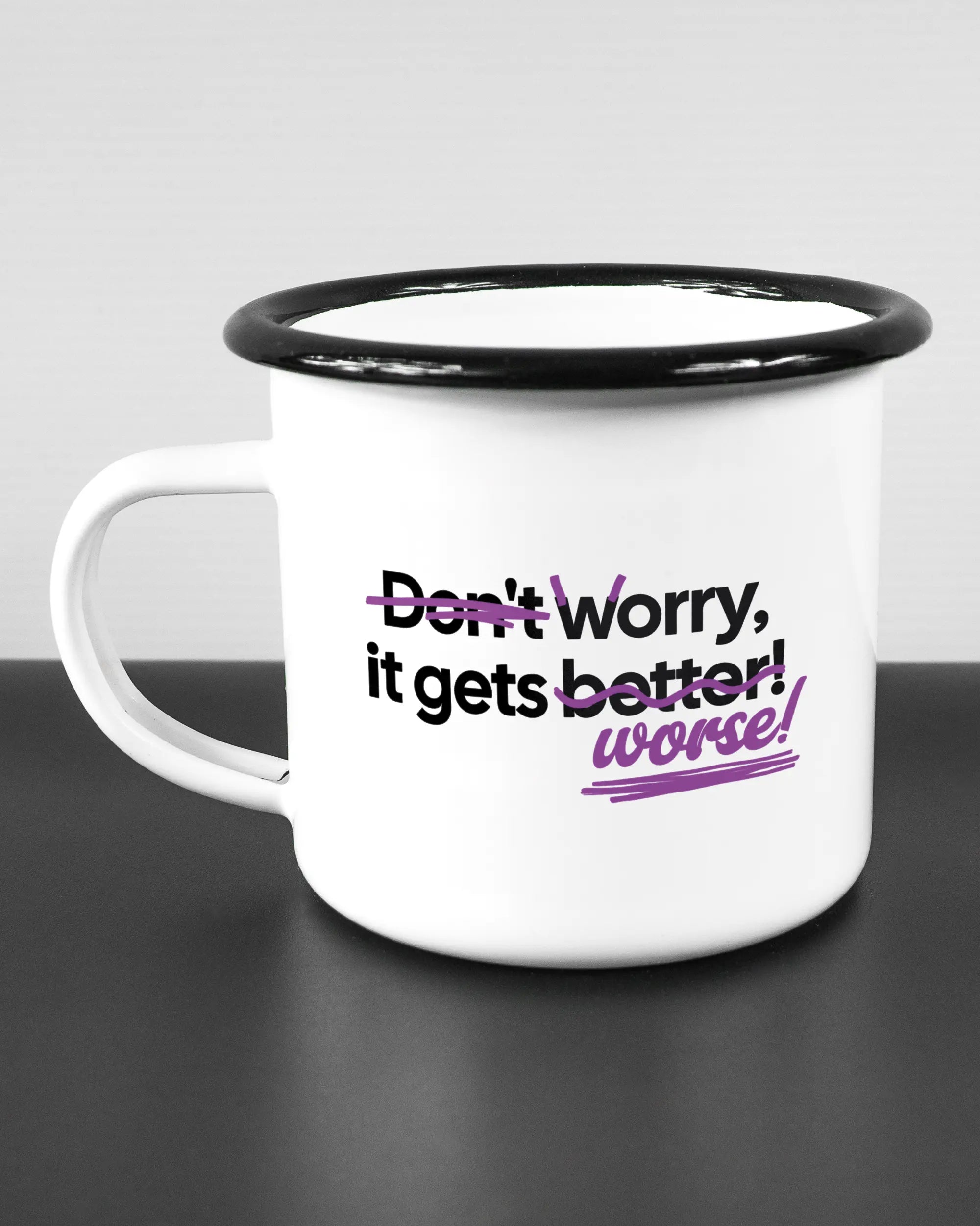 Titelbild, das die linke Seite der beidseitig bedruckten Emailletasse von Maondo mit dem Design Worry Worse in der Farbe White-Black auf einem Holztisch zeigt