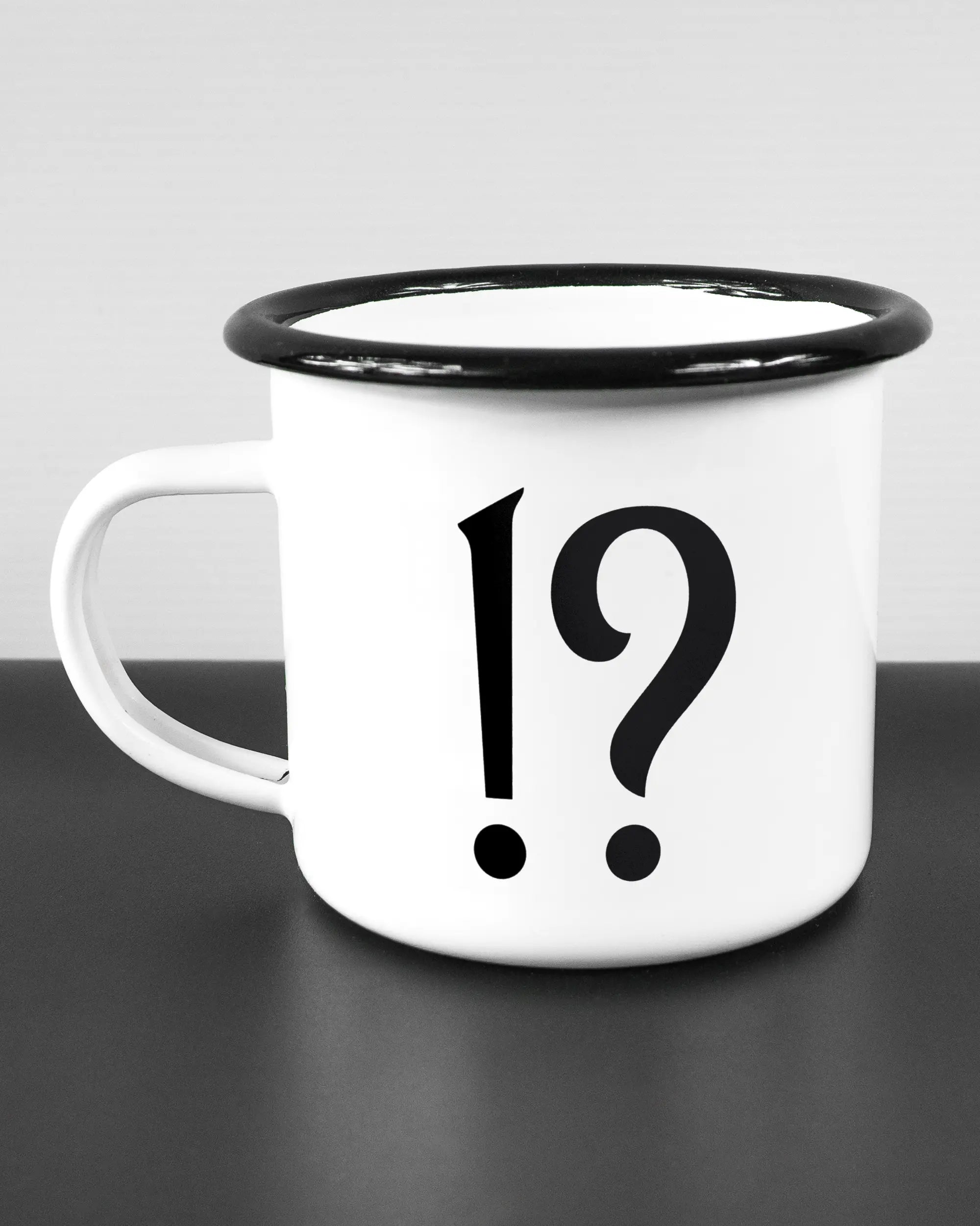 Titelbild, das die linke Seite der beidseitig bedruckten Emailletasse von Maondo mit dem Design WHAT in der Farbe White-Black auf einem Holztisch zeigt