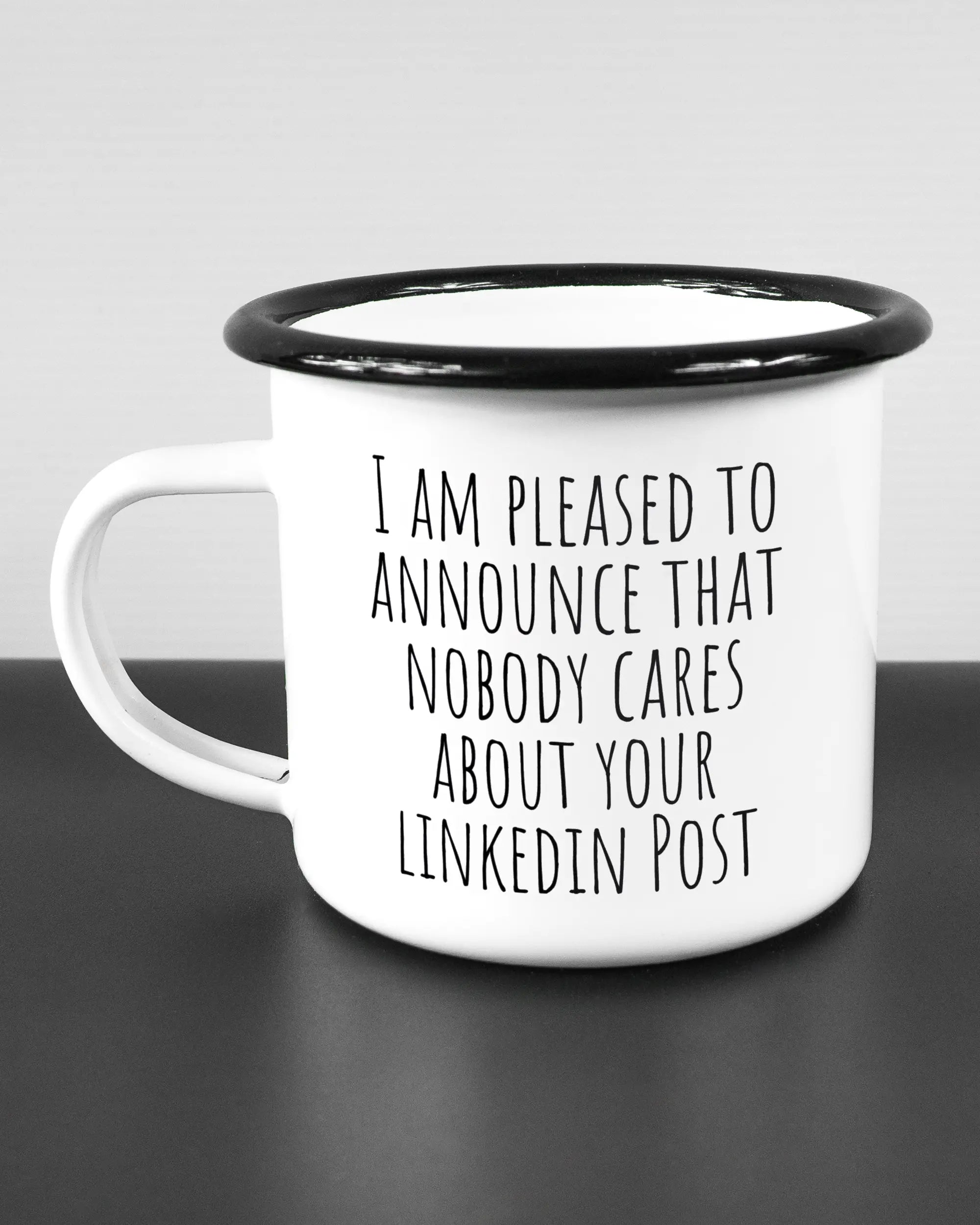 Titelbild, das die linke Seite der beidseitig bedruckten Emailletasse von Maondo mit dem Design Please Unannounce in der Farbe White-Black auf einem Holztisch zeigt