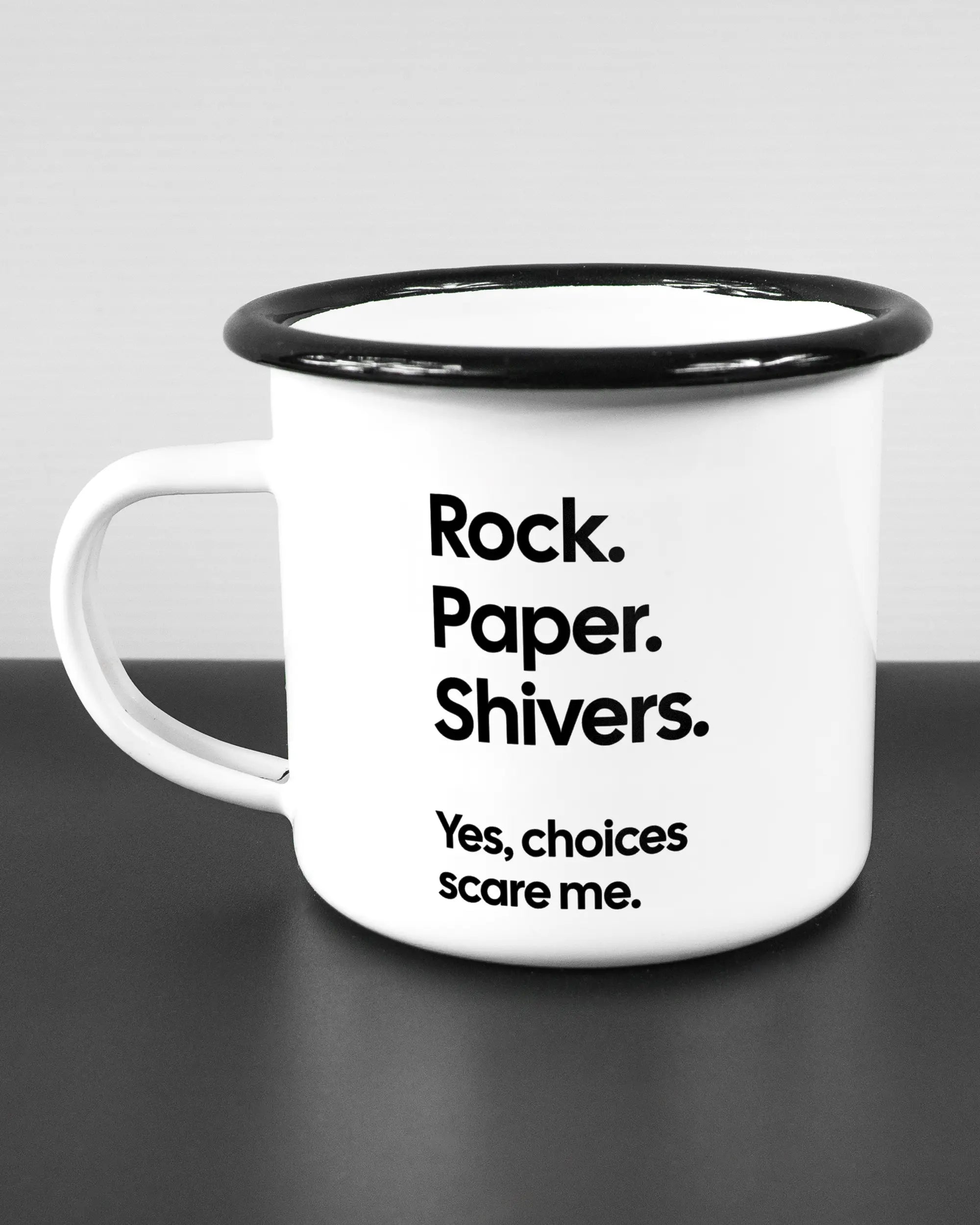 Titelbild, das die linke Seite der beidseitig bedruckten Emailletasse von Maondo mit dem Design Rock Paper Shivers in der Farbe White-Black auf einem Holztisch zeigt