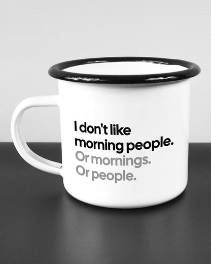 Titelbild, das die linke Seite der beidseitig bedruckten Emailletasse von Maondo mit dem Design Morning Person in der Farbe White-Black auf einem Holztisch zeigt