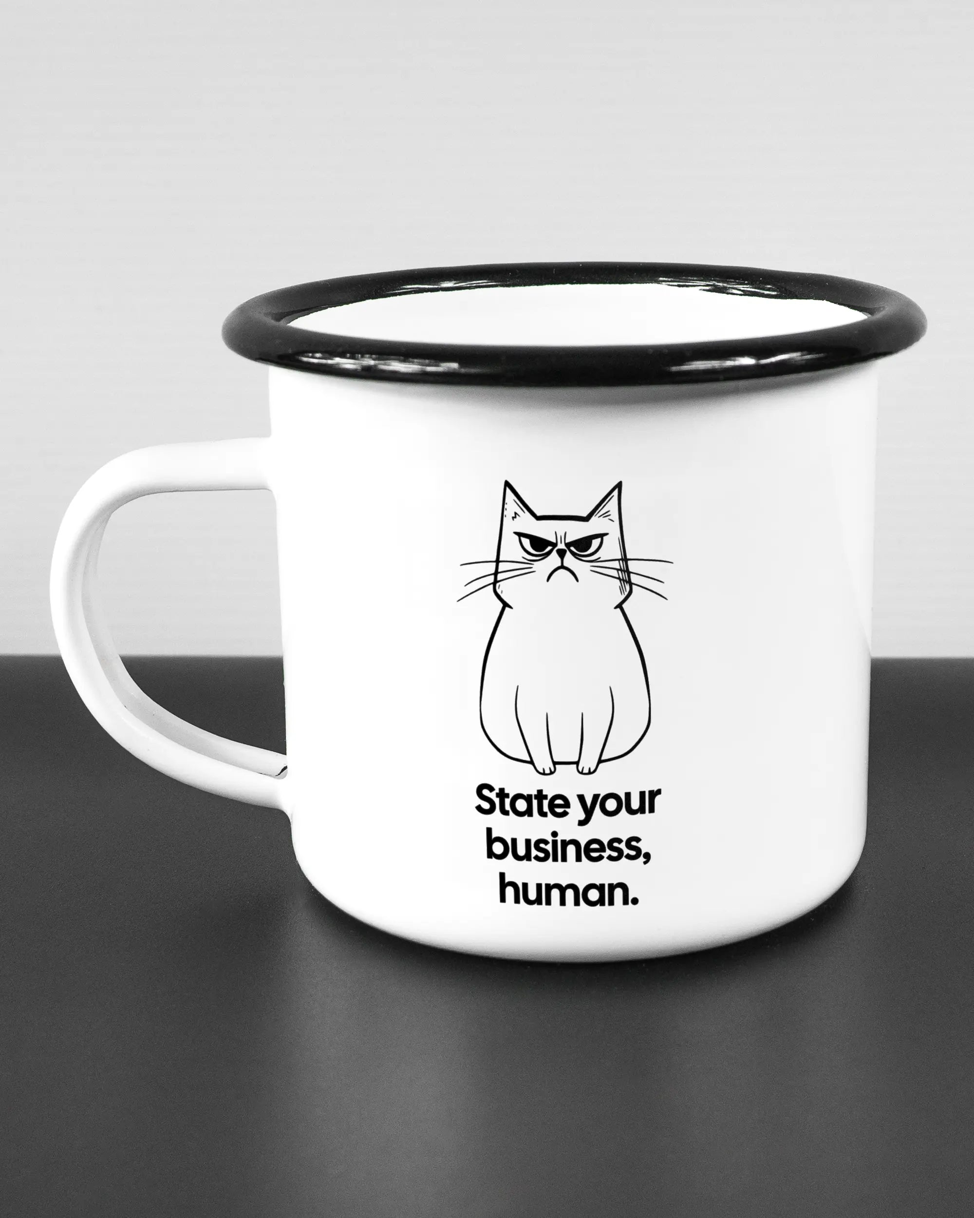 Titelbild, das die linke Seite der beidseitig bedruckten Emailletasse von Maondo mit dem Design Grumpy Purr in der Farbe White-Black auf einem Holztisch zeigt